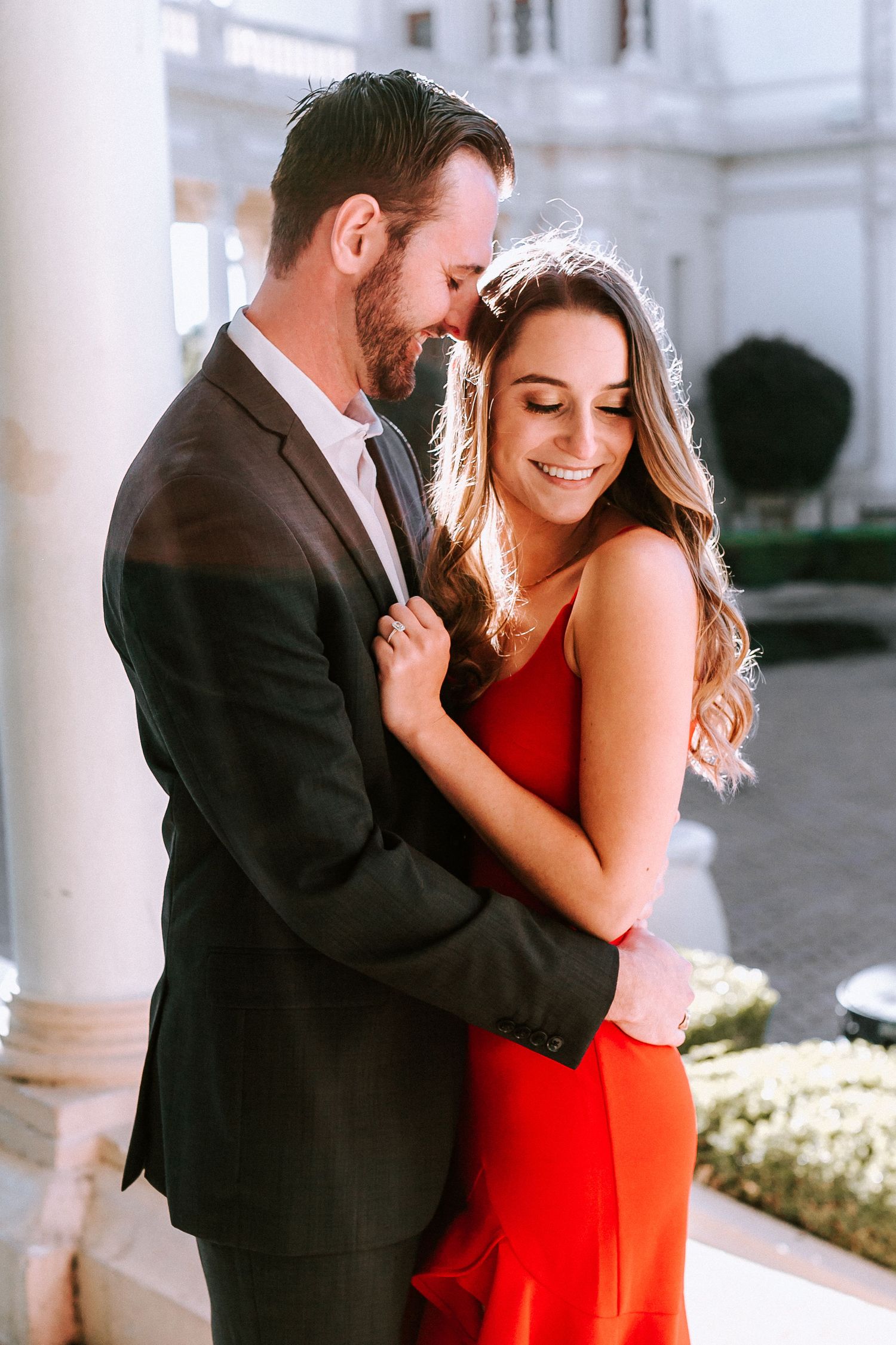Couple sharing a tender embrace outdoors, woman in red dress smiling joyfully.
