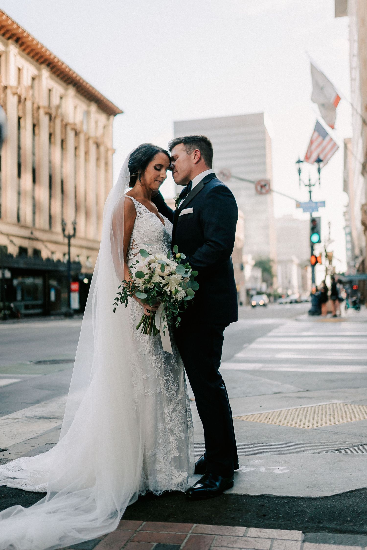 Bride and groom share romantic moment on urban street corner, bride in lace gown with veil holding white bouquet.