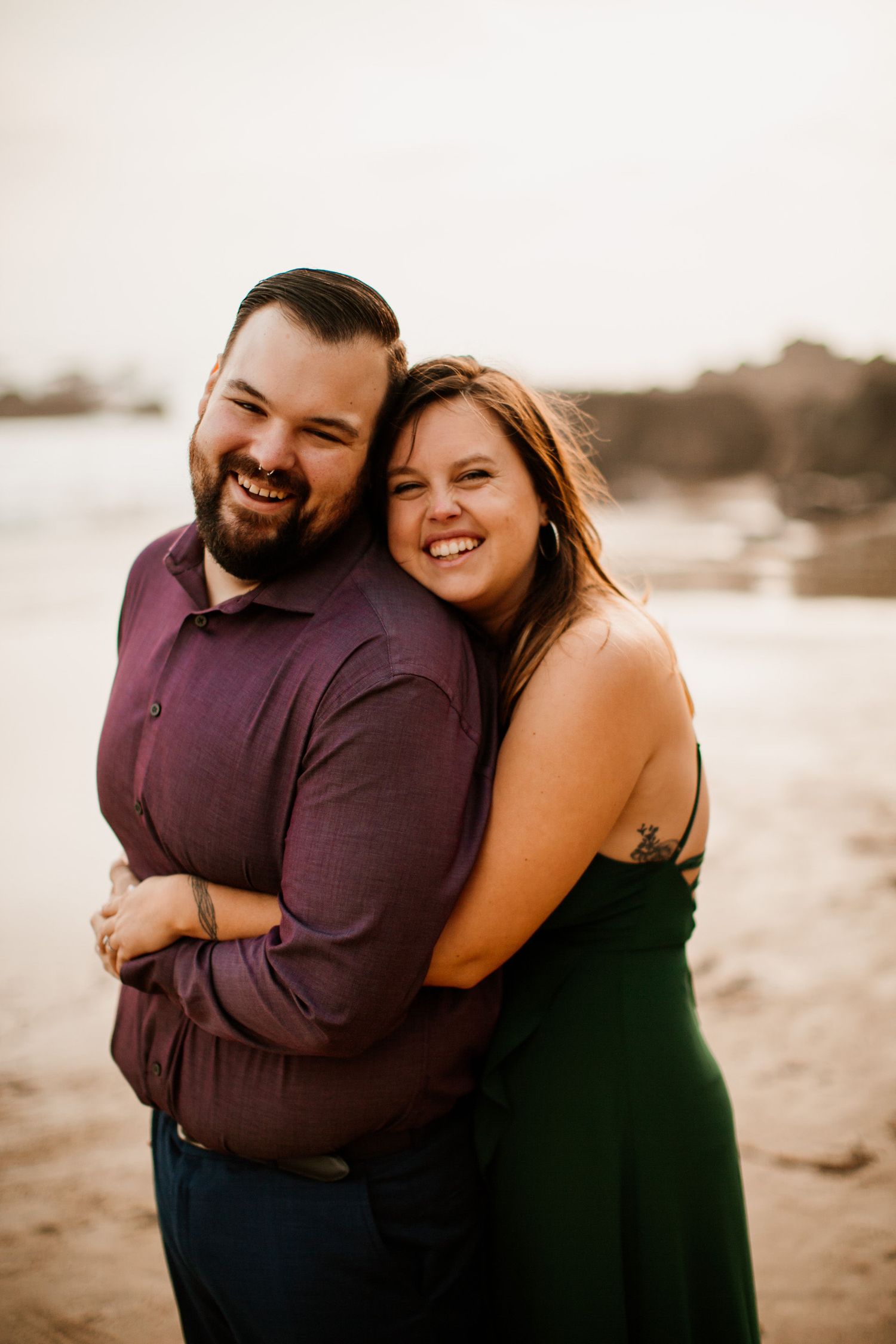 A smiling couple embraces on a sandy beach, man in purple shirt and woman in green dress, at golden hour.