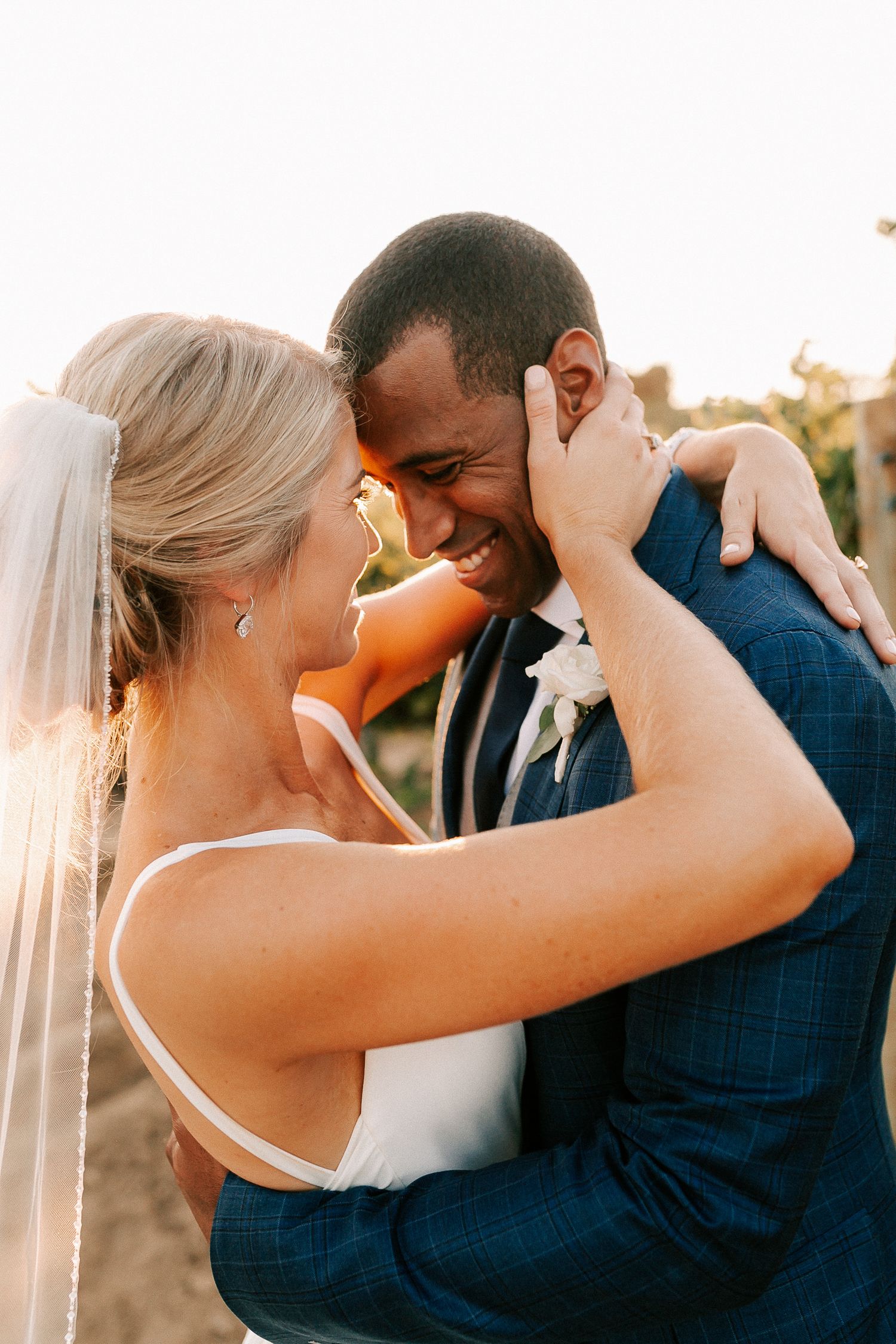 A joyful interracial couple shares an intimate embrace on their wedding day, with the bride in a veil and groom in a navy suit.