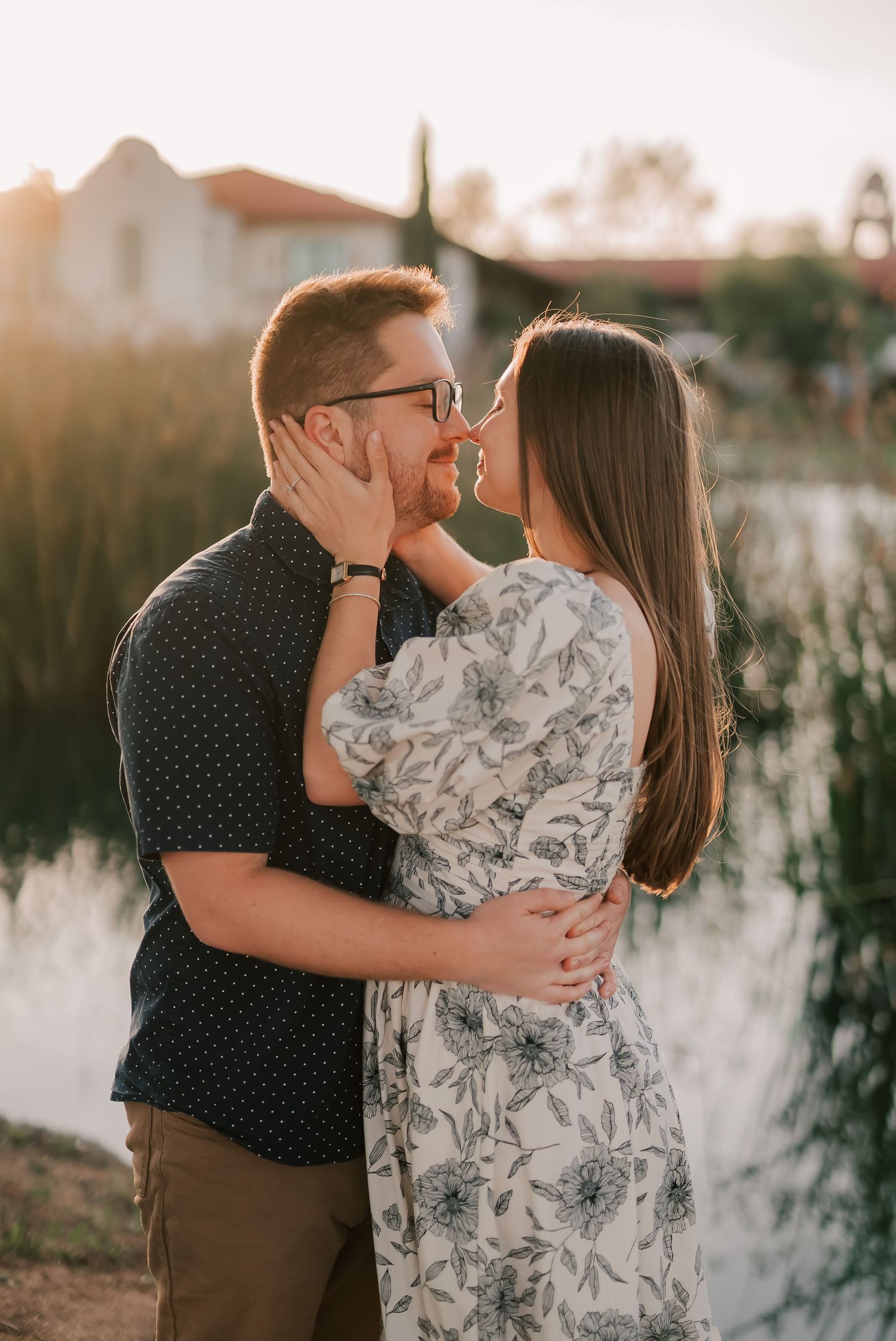 Smiling couple embracing and touching noses at golden hour beside a calm pond, the woman in a floral dress and the man in glasses.