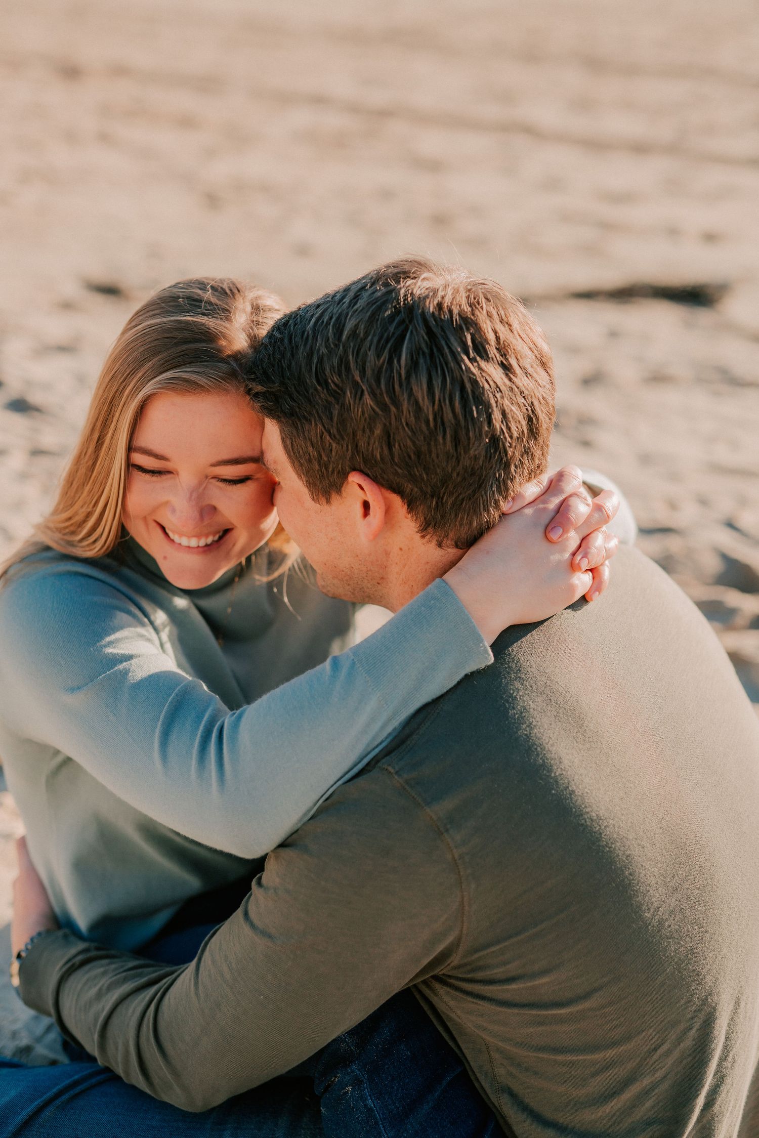 Joyful couple cuddling on sandy beach in matching green sweaters in warm sunlight.