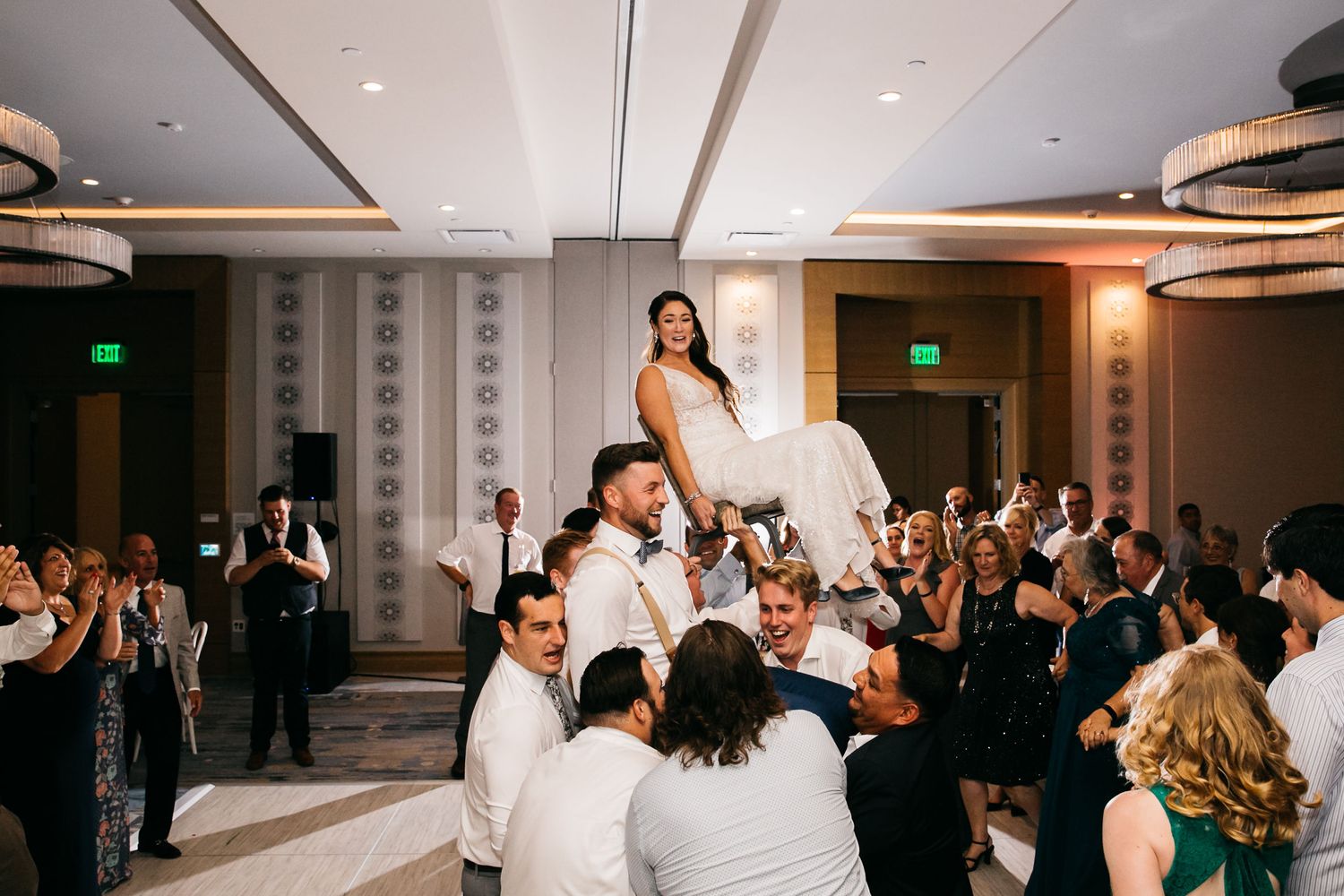 Bride lifted by groom and groomsmen on wedding dance floor surrounded by cheering guests at elegant hotel reception.