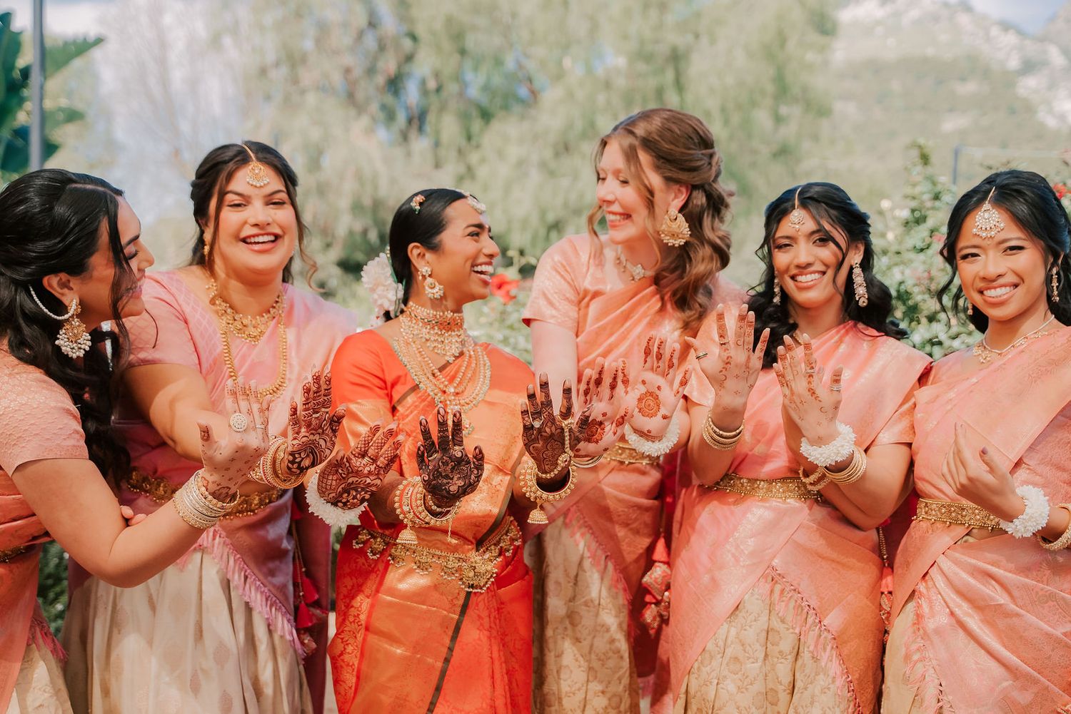 Bridesmaids in pink and orange sarees joyfully displaying mehndi henna designs at Indian wedding.
