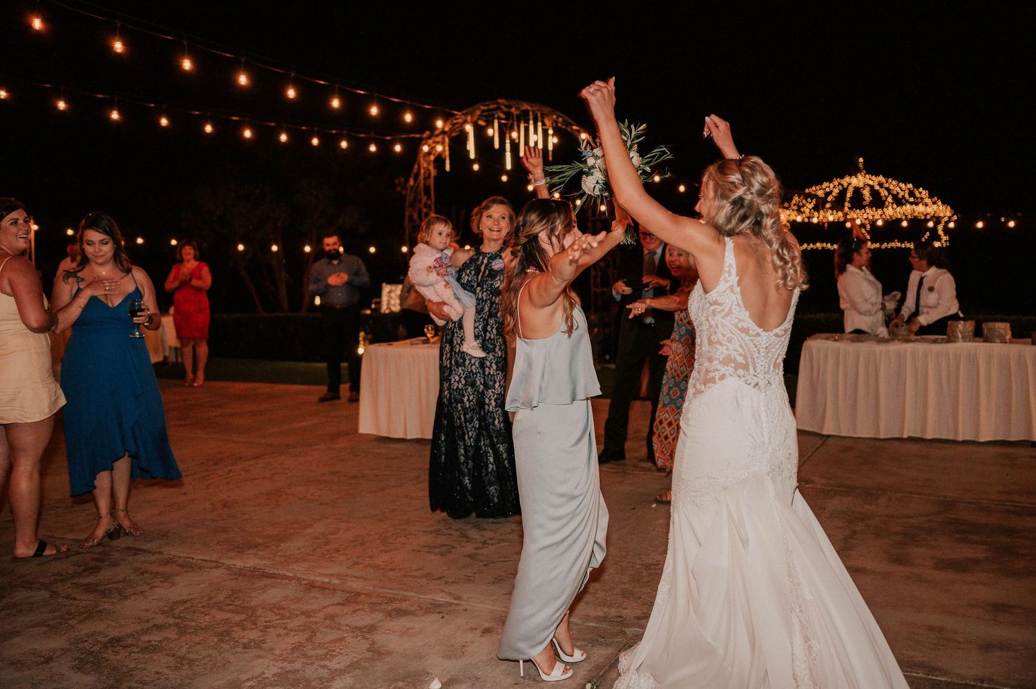 Bride in white lace gown tosses bouquet to guests at outdoor wedding reception with string lights.