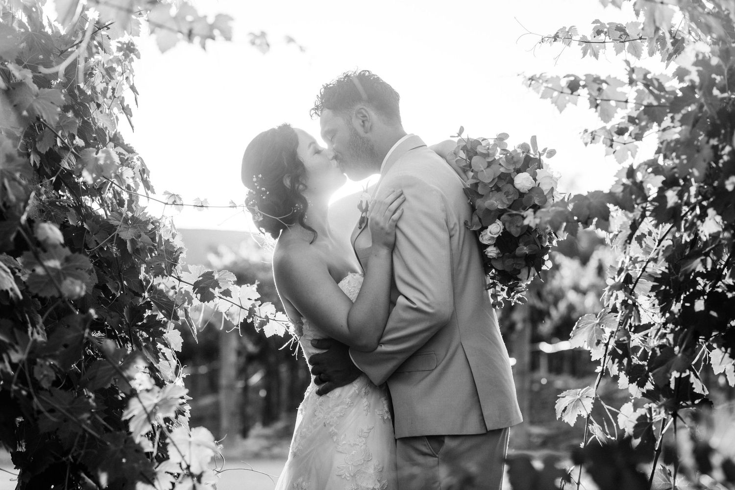 Black and white photo of a couple sharing a kiss at their wedding ceremony surrounded by lush greenery and floral arrangements.