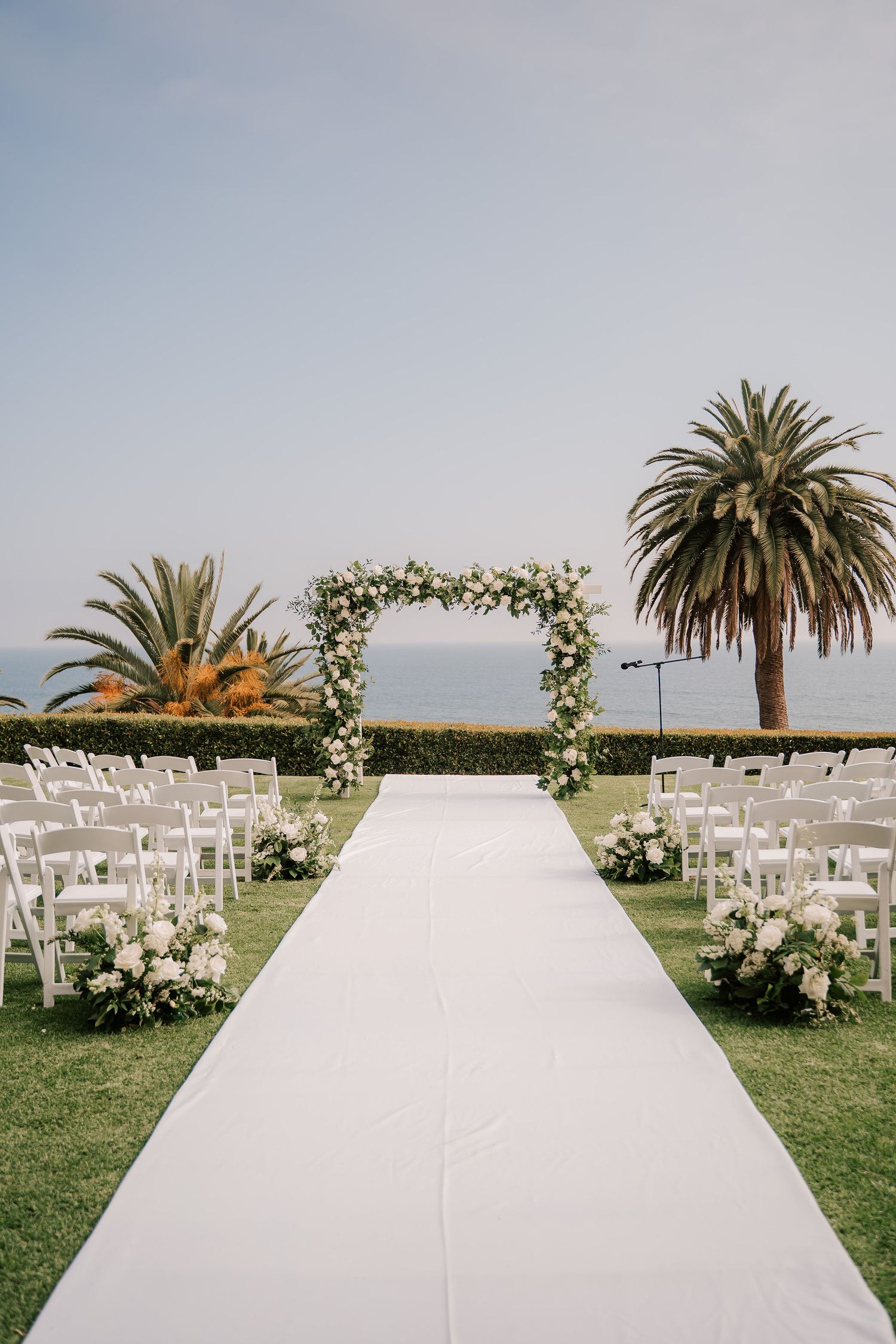 White aisle runner leads to a floral arch at an oceanfront wedding ceremony with white chairs, palm trees and flower arrangements.