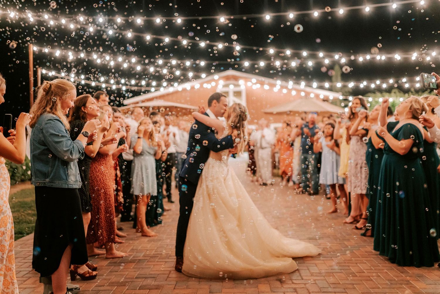 Bride and groom share first dance under string lights surrounded by guests blowing bubbles at outdoor wedding reception.