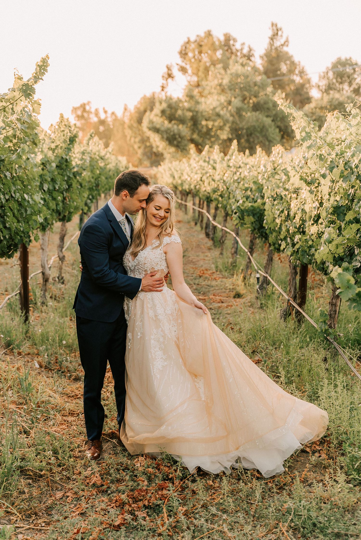 Couple embracing in a sunlit vineyard, bride in a flowing champagne lace gown, groom in a navy suit.