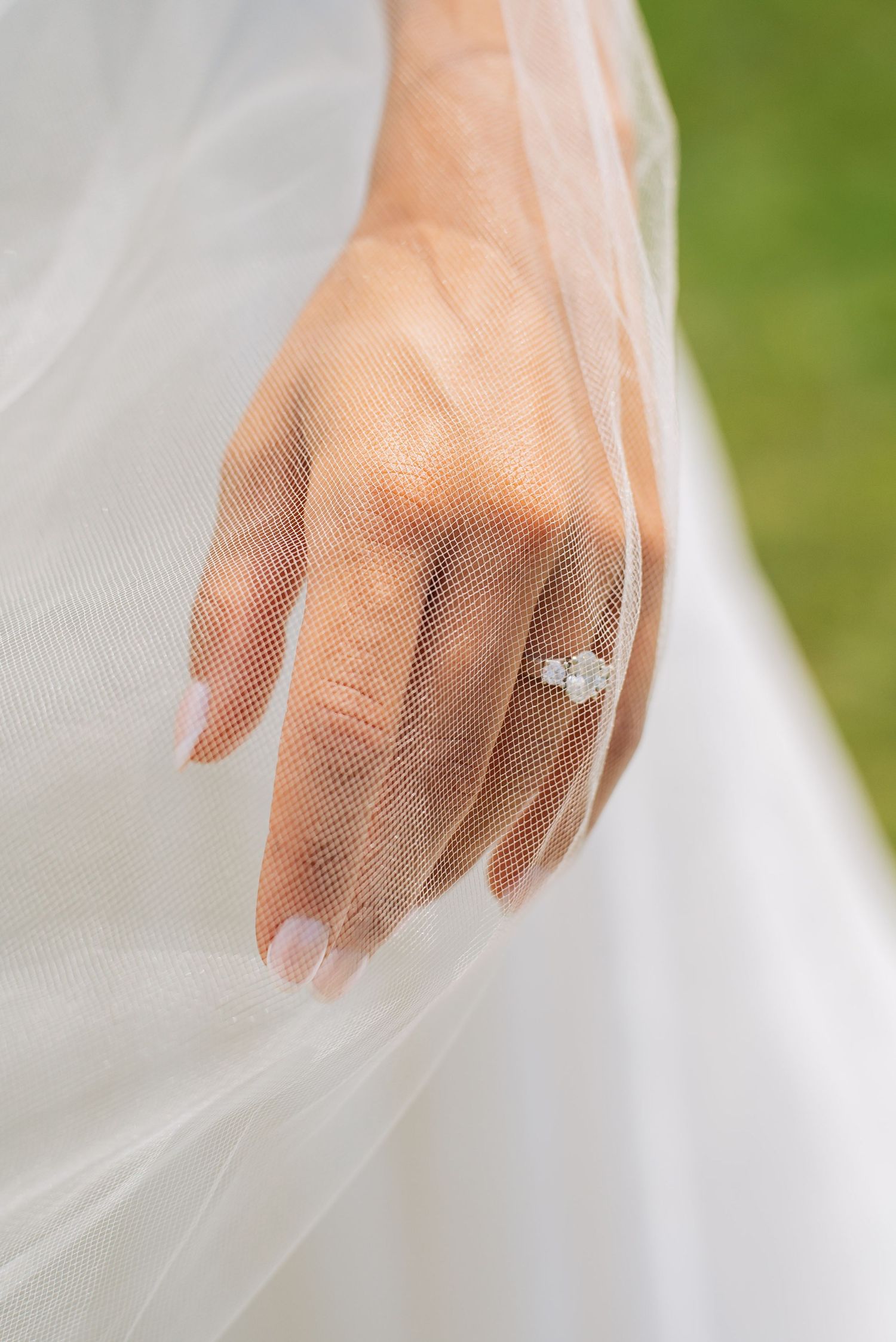 Close-up of a bride's hand wearing a diamond engagement ring, draped with sheer white wedding veil fabric.