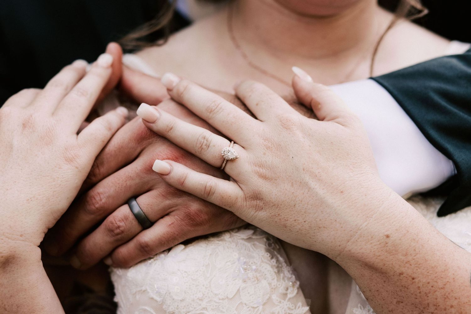 Multiple hands stacked together showing wedding bands on bride in white lace dress at wedding ceremony.