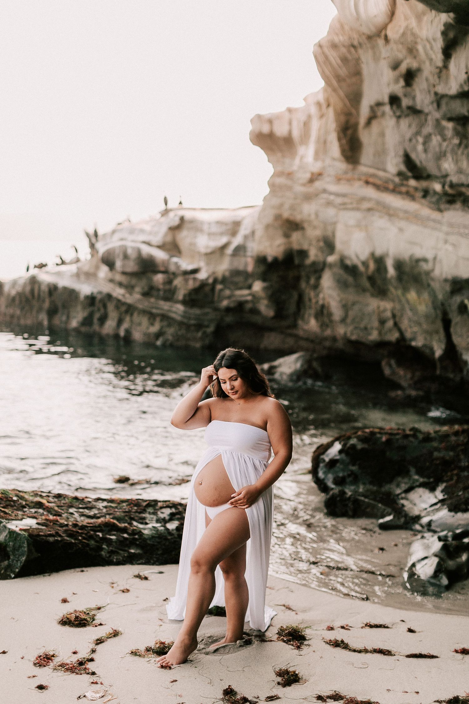 Pregnant woman in white strapless maternity dress posing on beach with rocky cliffs in background.