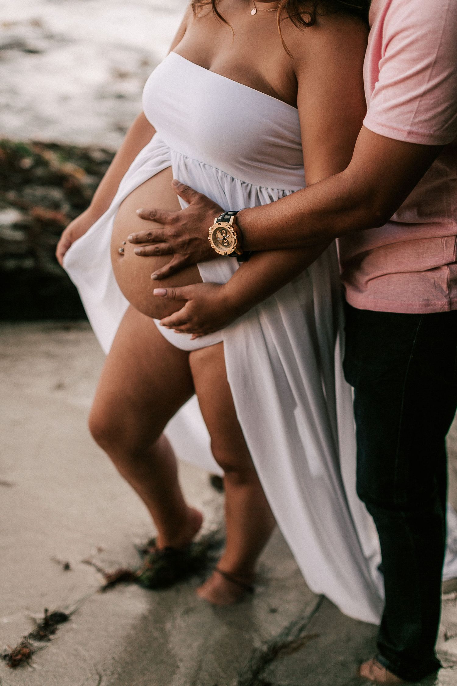 Close-up of pregnant belly with partner embracing woman in white maternity dress on beach.