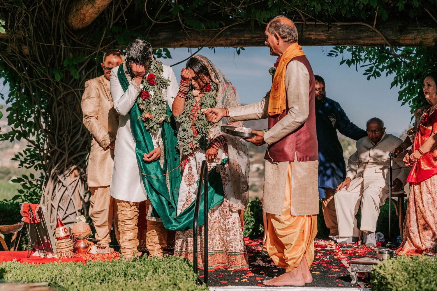 Officiant presents sacred items to couple during traditional Indian wedding ceremony under tree canopy.