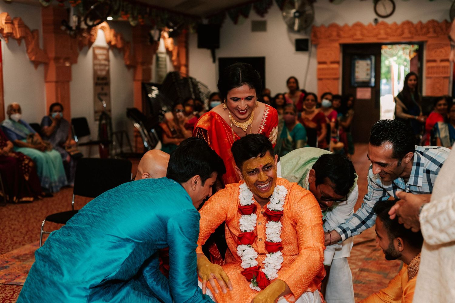 Groom in orange kurta surrounded by laughing family and friends during a joyful Indian wedding celebration.