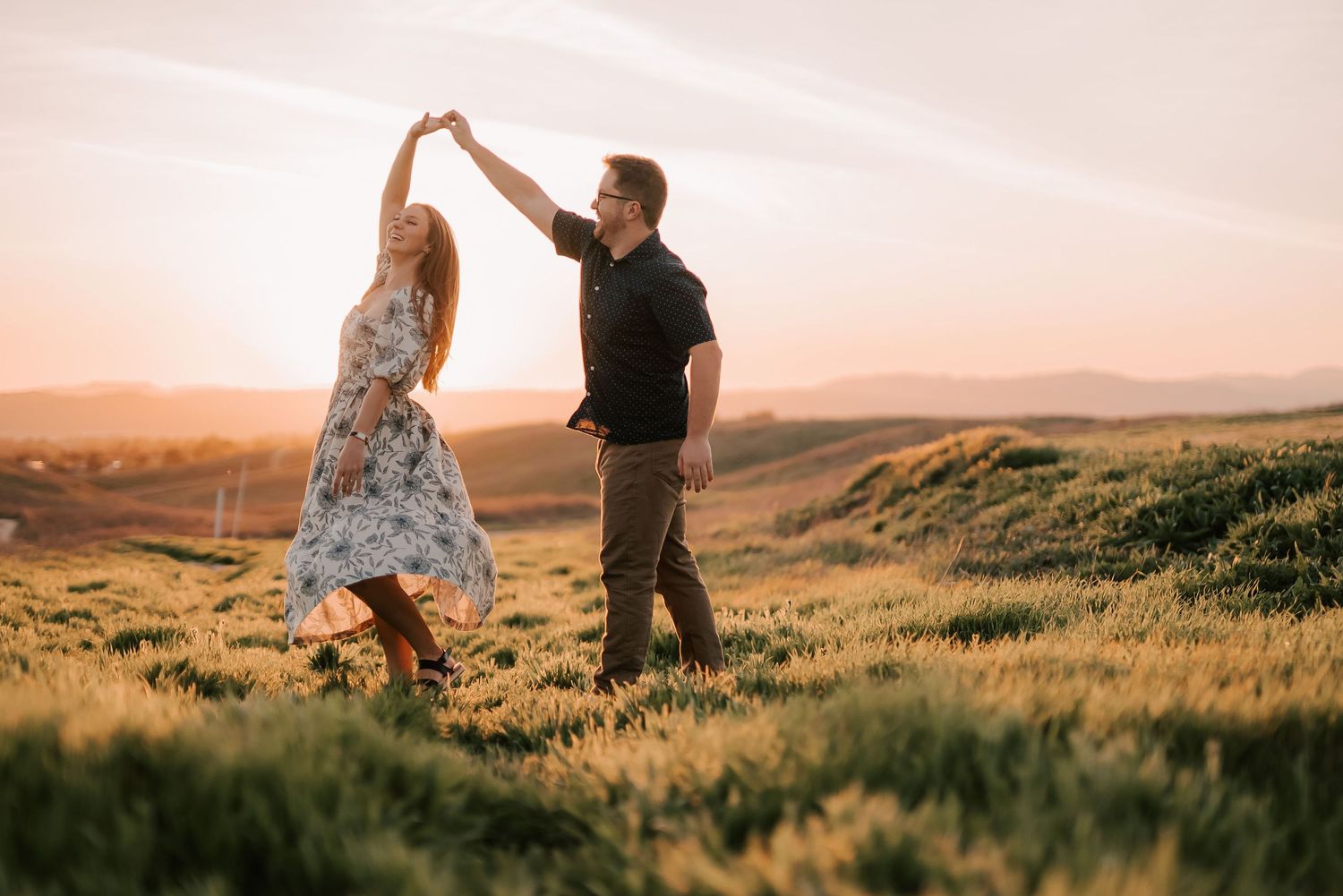 Couple dancing at sunset in a golden field, man spinning woman in floral dress with rolling hills backdrop.