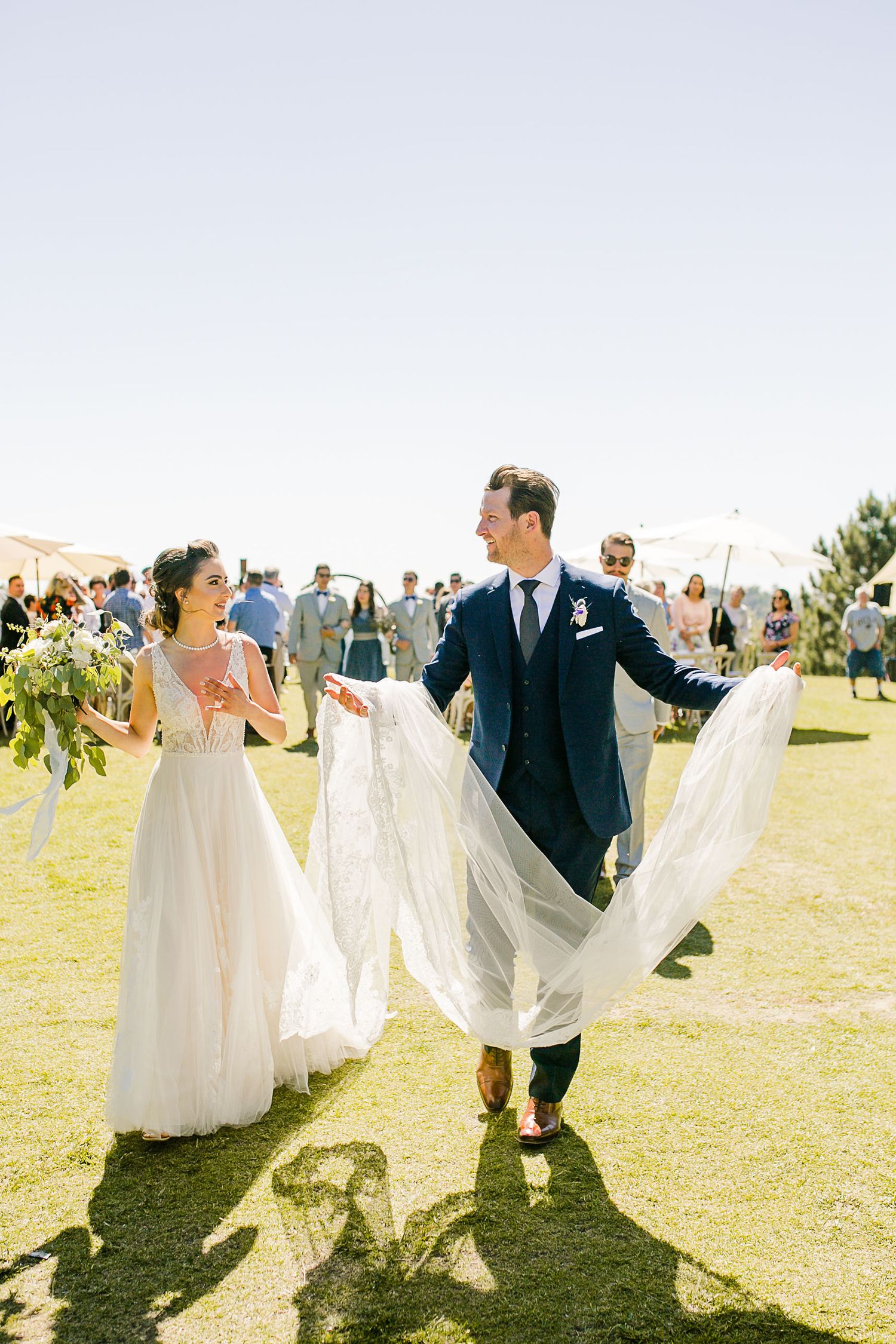 Bride and groom walk joyfully down the aisle outdoors on a sunny day, groom holding the wedding dress train.
