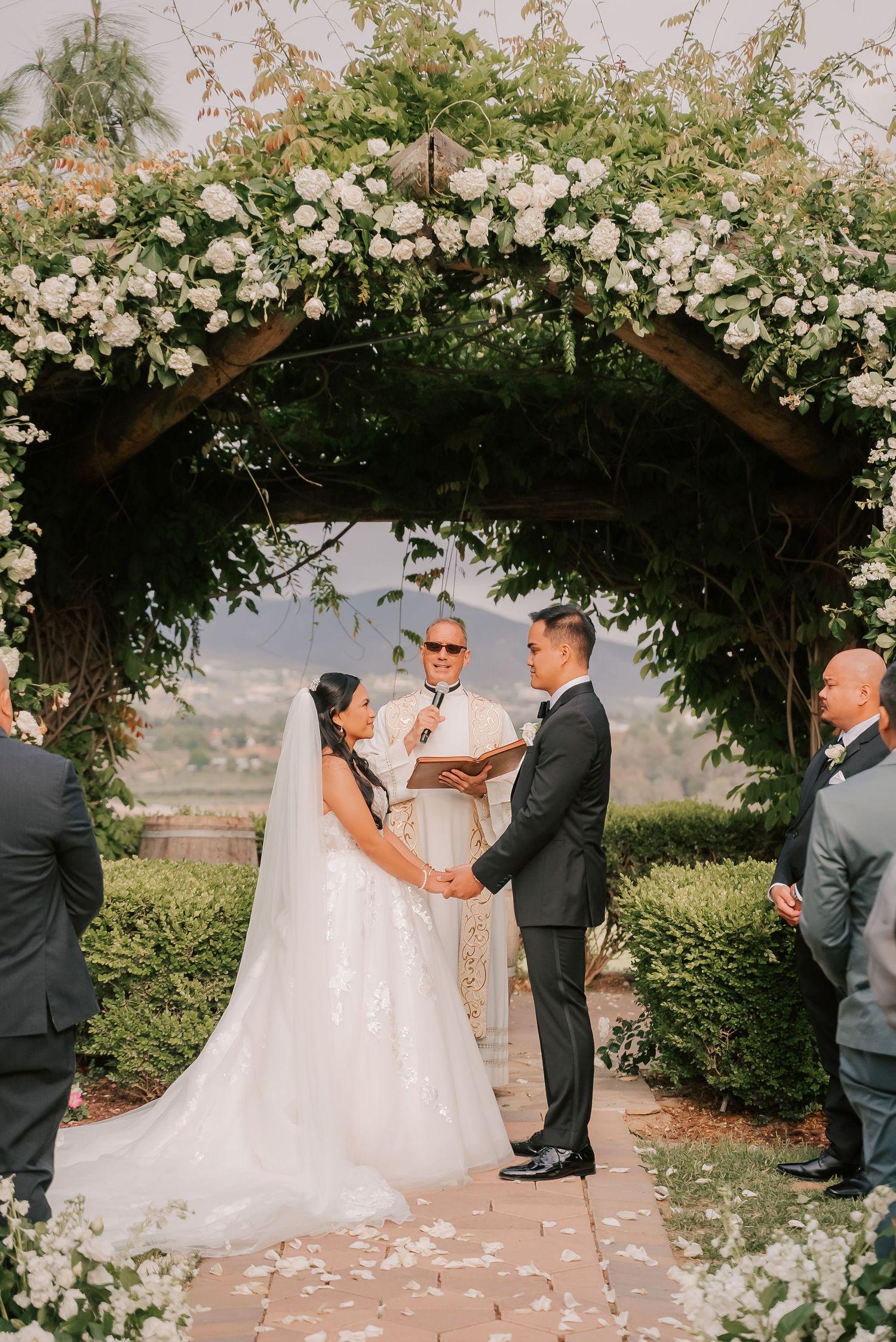Bride and groom exchange vows under a floral arch adorned with white roses and greenery at an outdoor wedding ceremony.