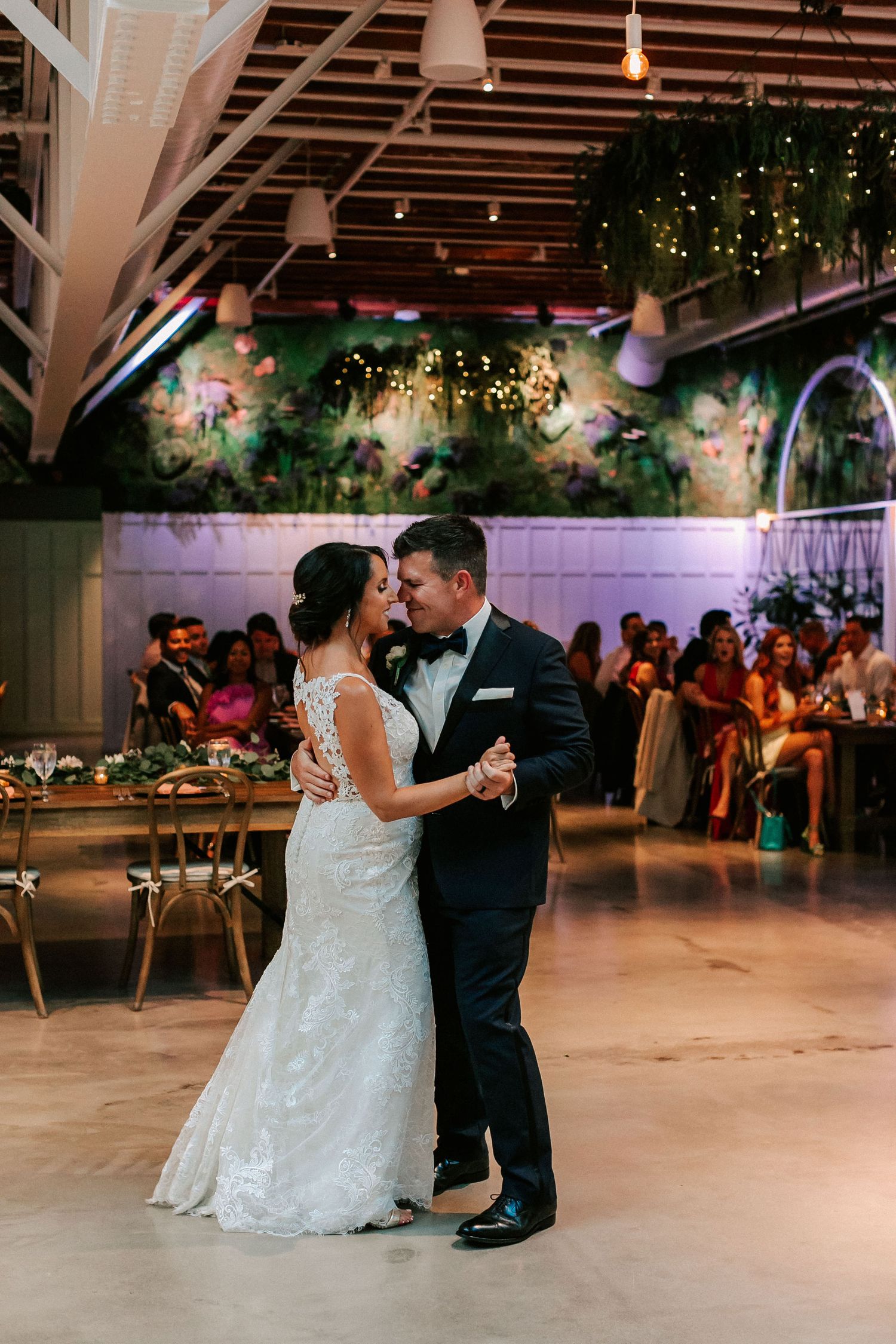 Bride and groom share their first dance in an elegant wedding venue with string lights and lush greenery backdrop.