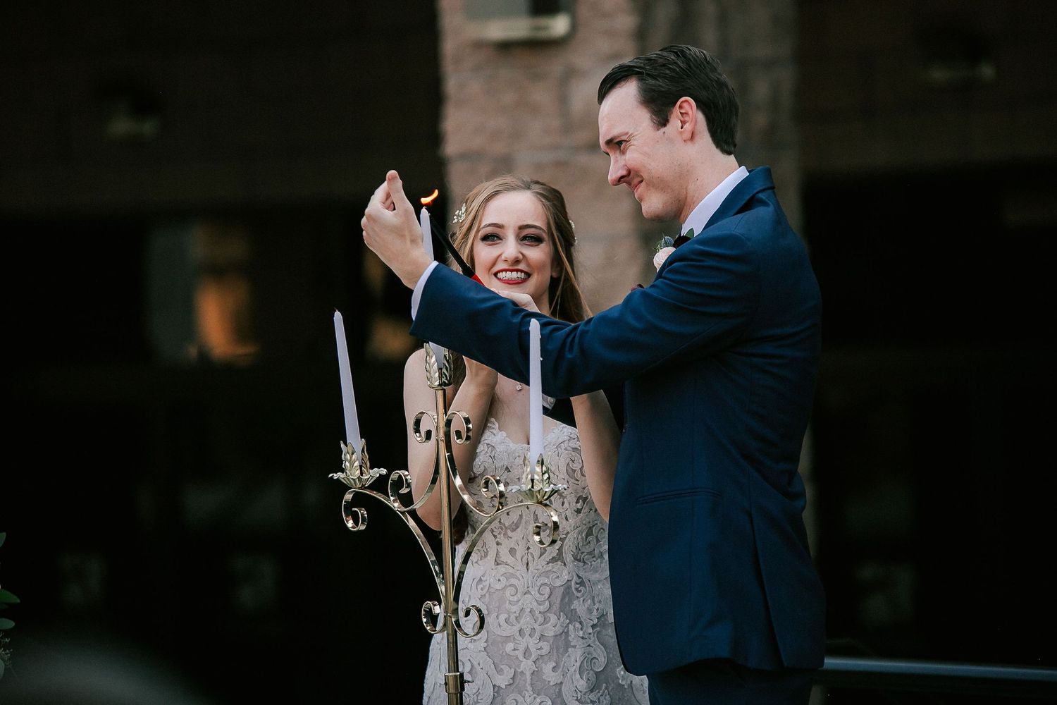 Bride and groom lighting candles together outdoors during wedding ceremony, smiling joyfully.