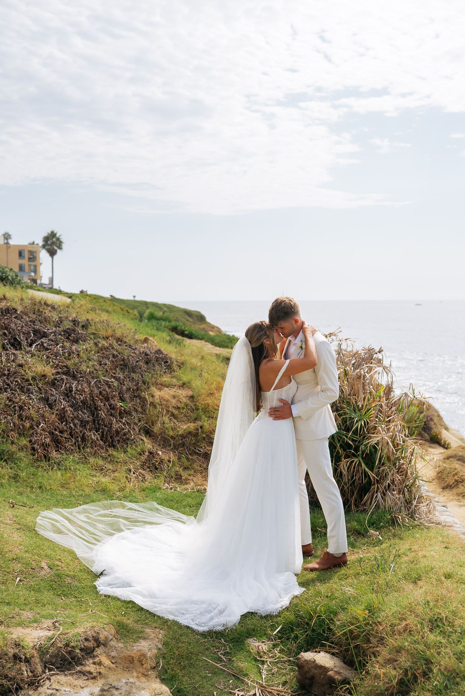 Bride and groom share a romantic kiss on a coastal cliffside, with the bride's long veil flowing in the breeze.