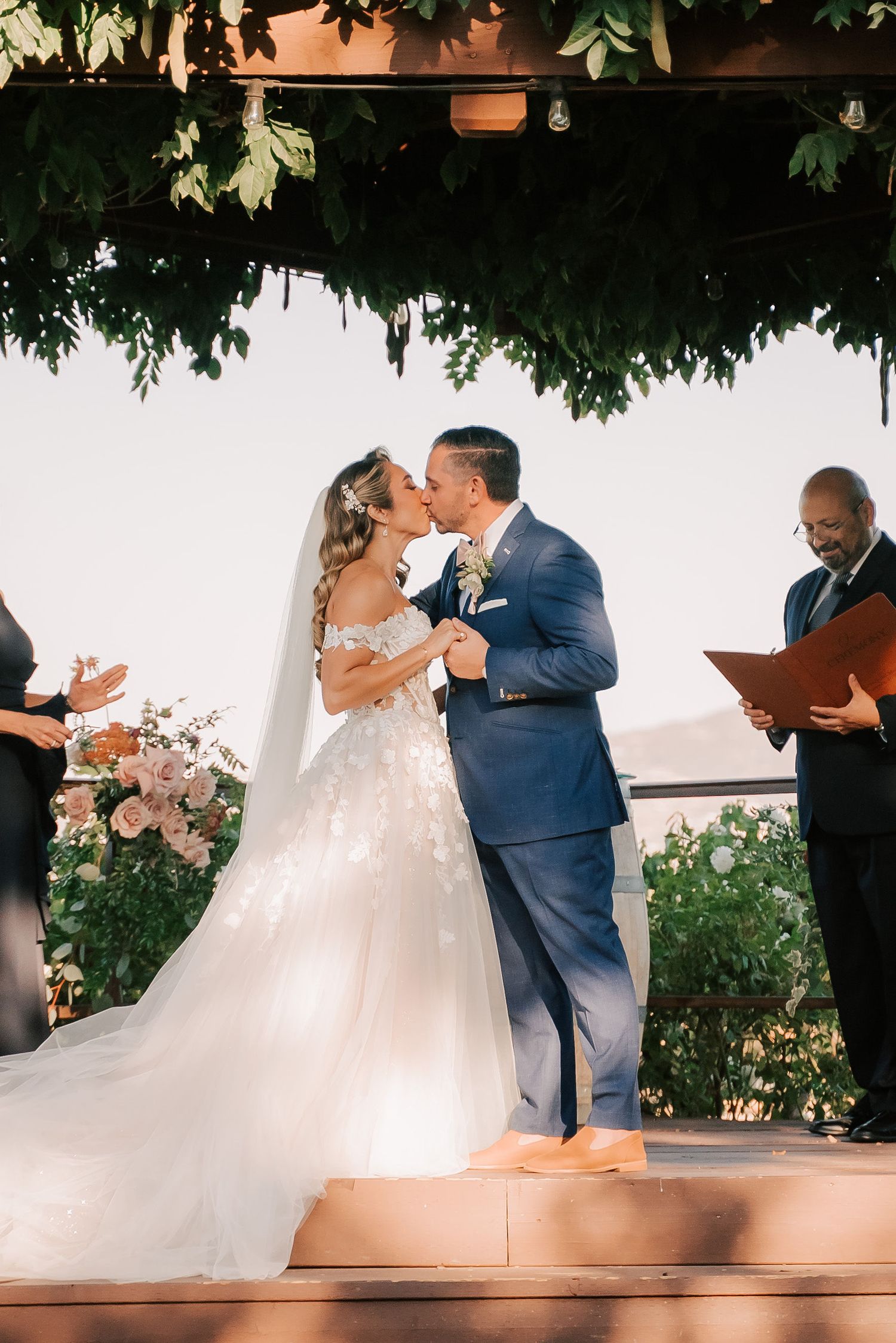 Bride and groom share first kiss at outdoor wedding ceremony under lush greenery arbor, officiant holding book nearby.
