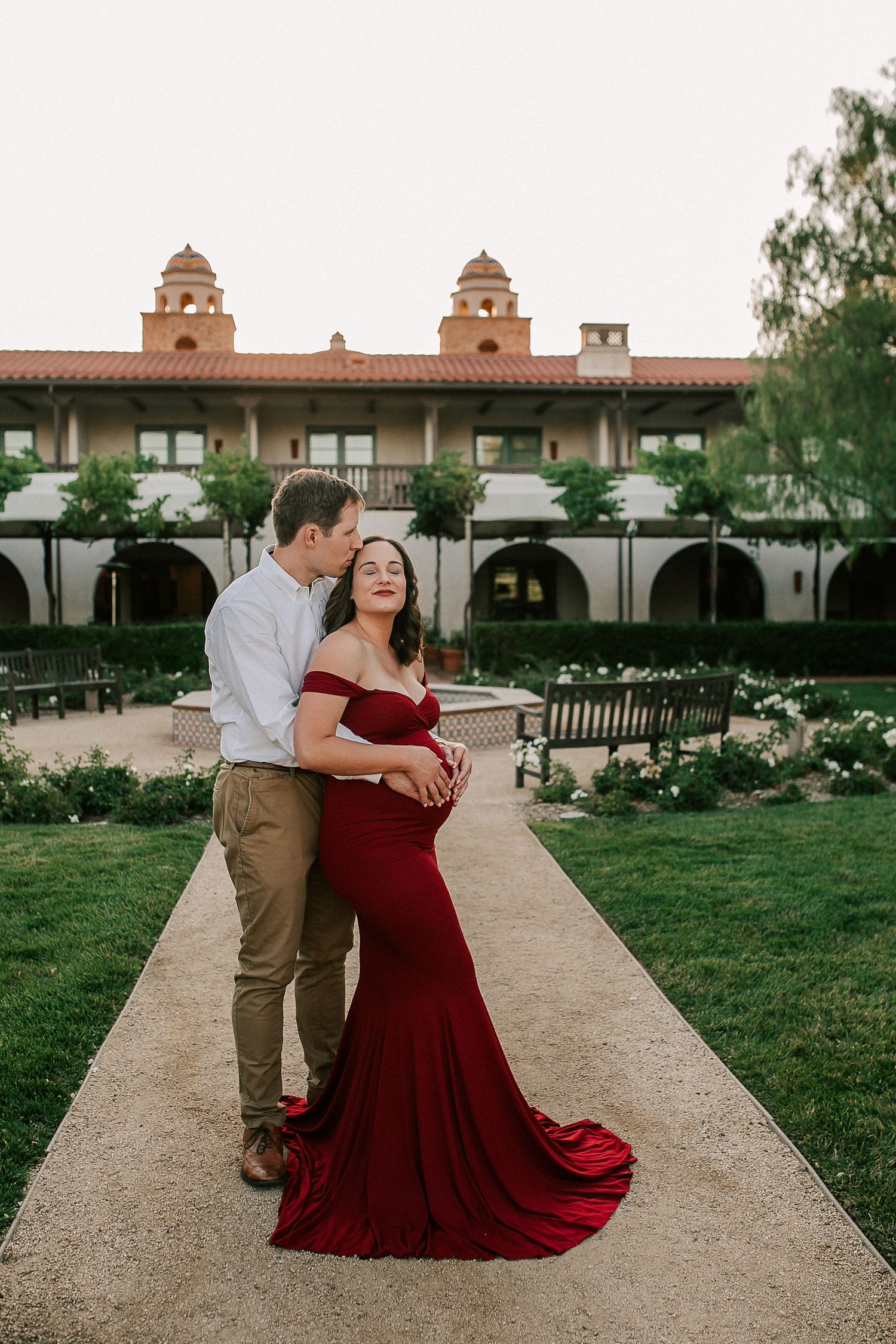 Couple poses in courtyard, pregnant woman in red gown, man in white shirt embracing her waist.