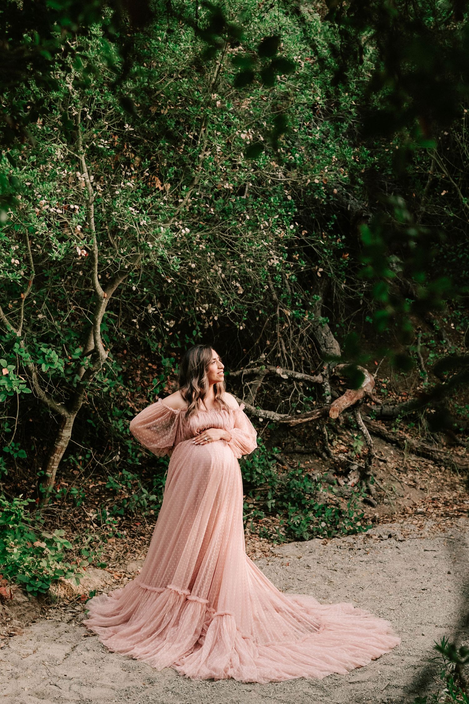 Pregnant woman in flowing pink tulle gown poses outdoors among lush green trees in elegant maternity photo.