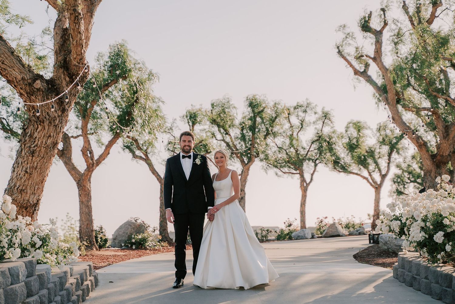 Bride and groom walking together outdoors at a wedding venue surrounded by olive trees and white floral arrangements.