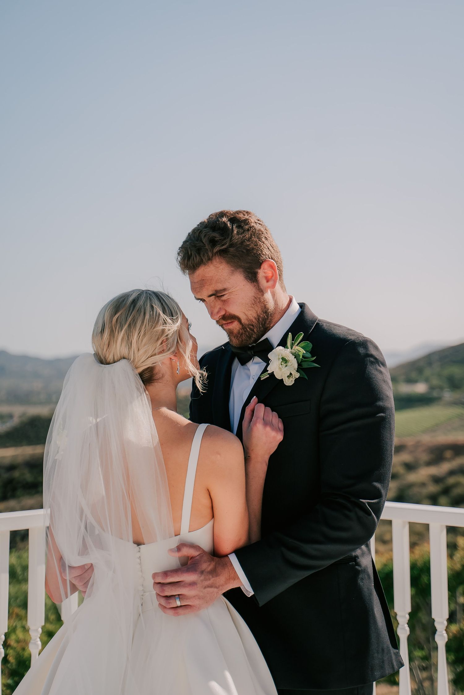 Bride and groom share an intimate moment on a balcony with scenic hills, she in a white gown with veil, he in a black tuxedo.