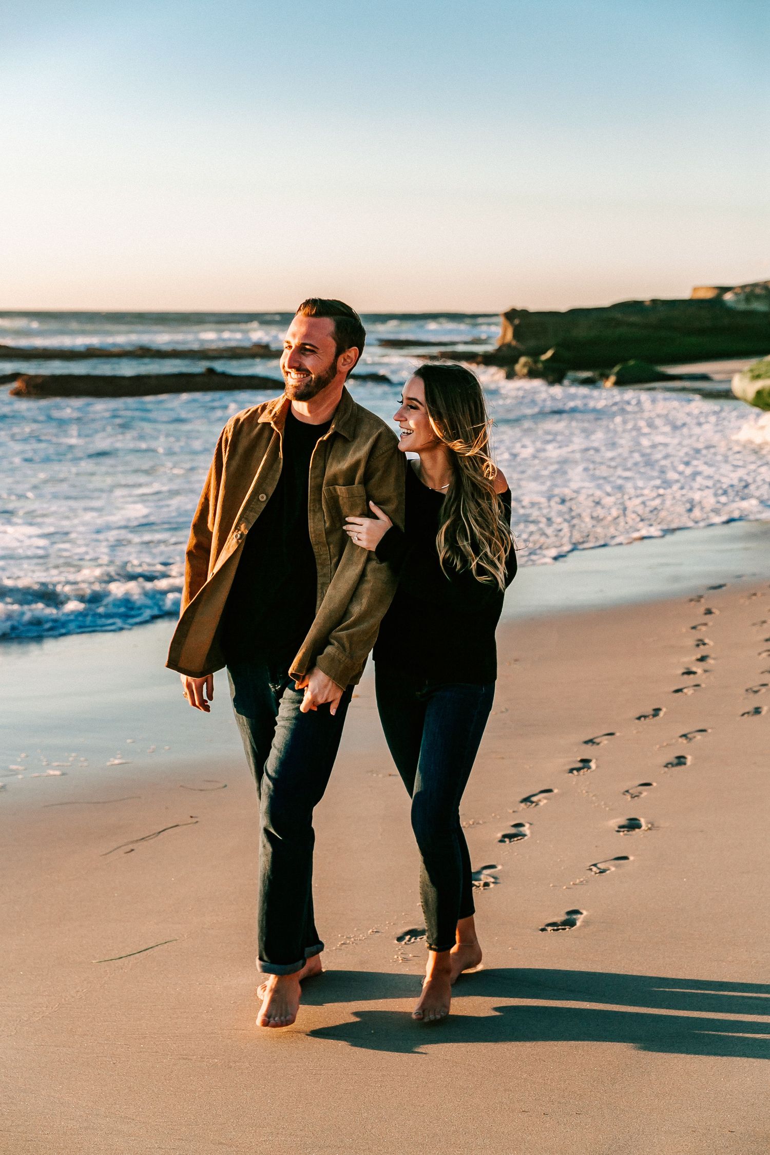 Barefoot couple walking hand in hand along shoreline at sunset, leaving footprints in sand.