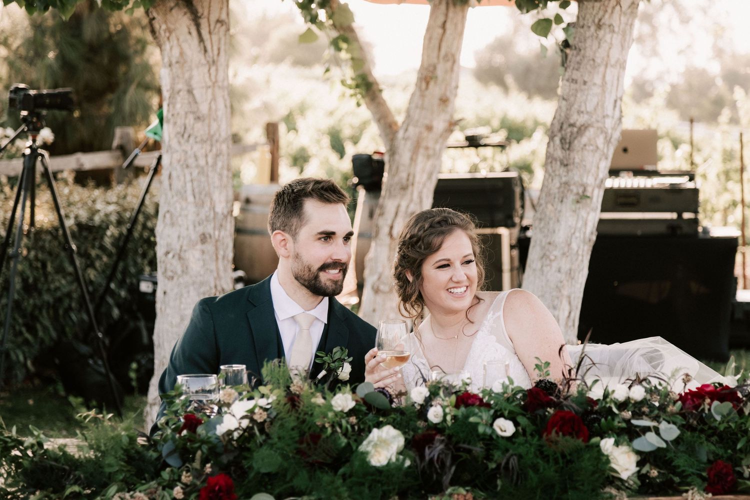 Bride and groom smiling at outdoor wedding reception table adorned with red and white floral arrangements.