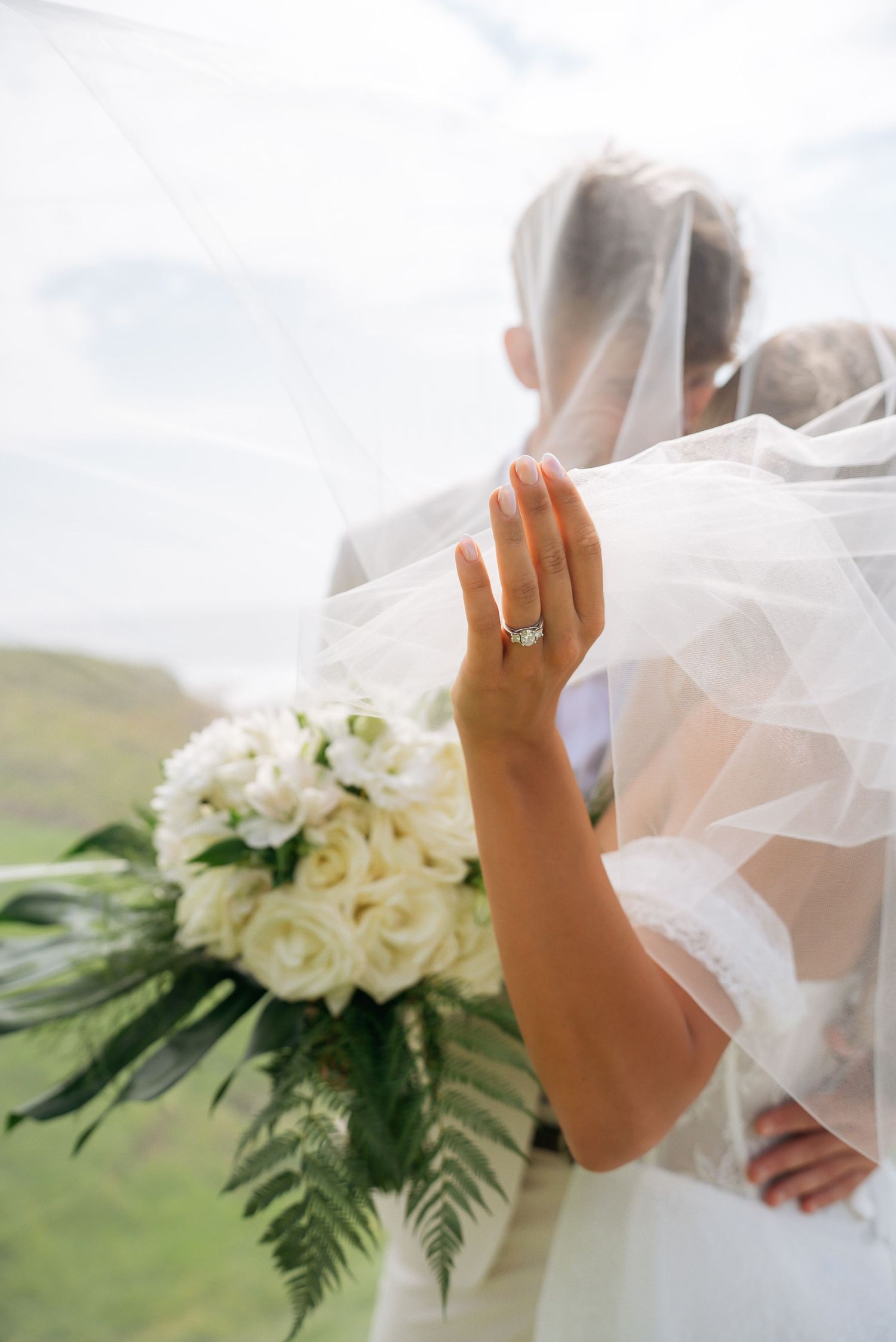 Bride holding white rose bouquet with engagement ring visible, veil flowing, embracing groom outdoors at wedding.