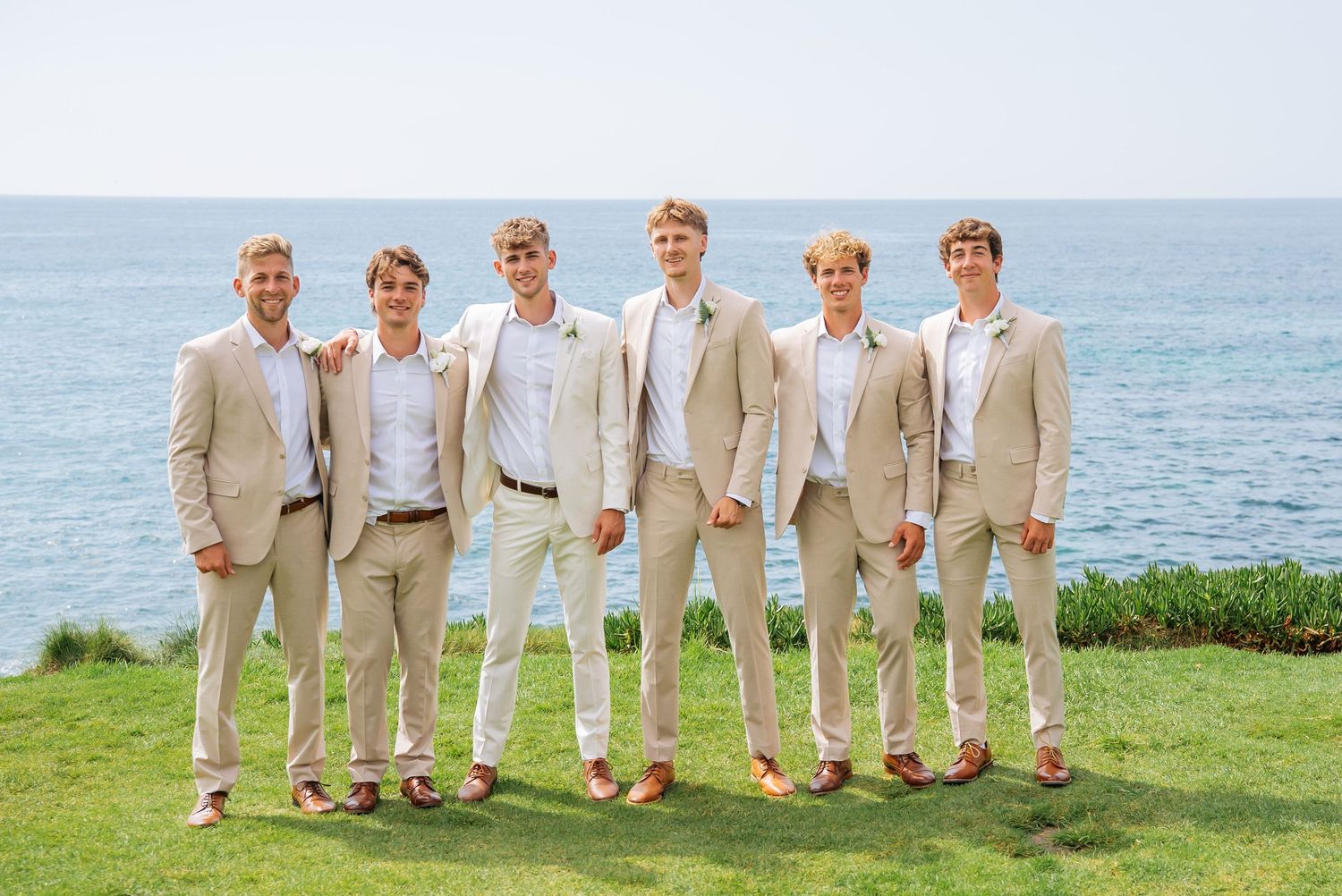 Six groomsmen in matching tan suits pose together on a green lawn with a scenic ocean backdrop.