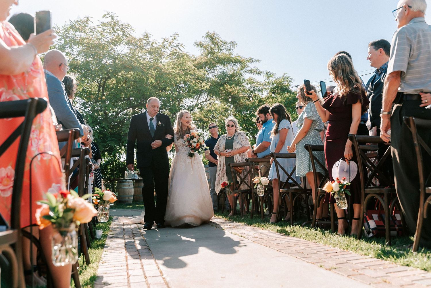 Bride walks down the aisle with her father at an outdoor wedding ceremony, guests standing on both sides.