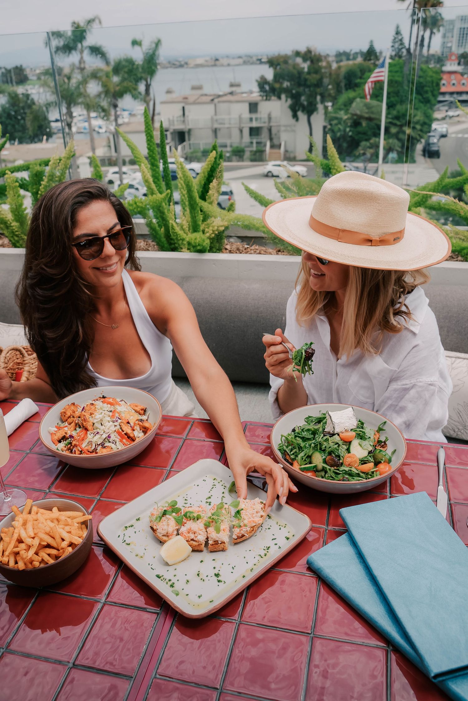 Two women dining al fresco on a rooftop patio, enjoying pasta and salad dishes with an ocean view.