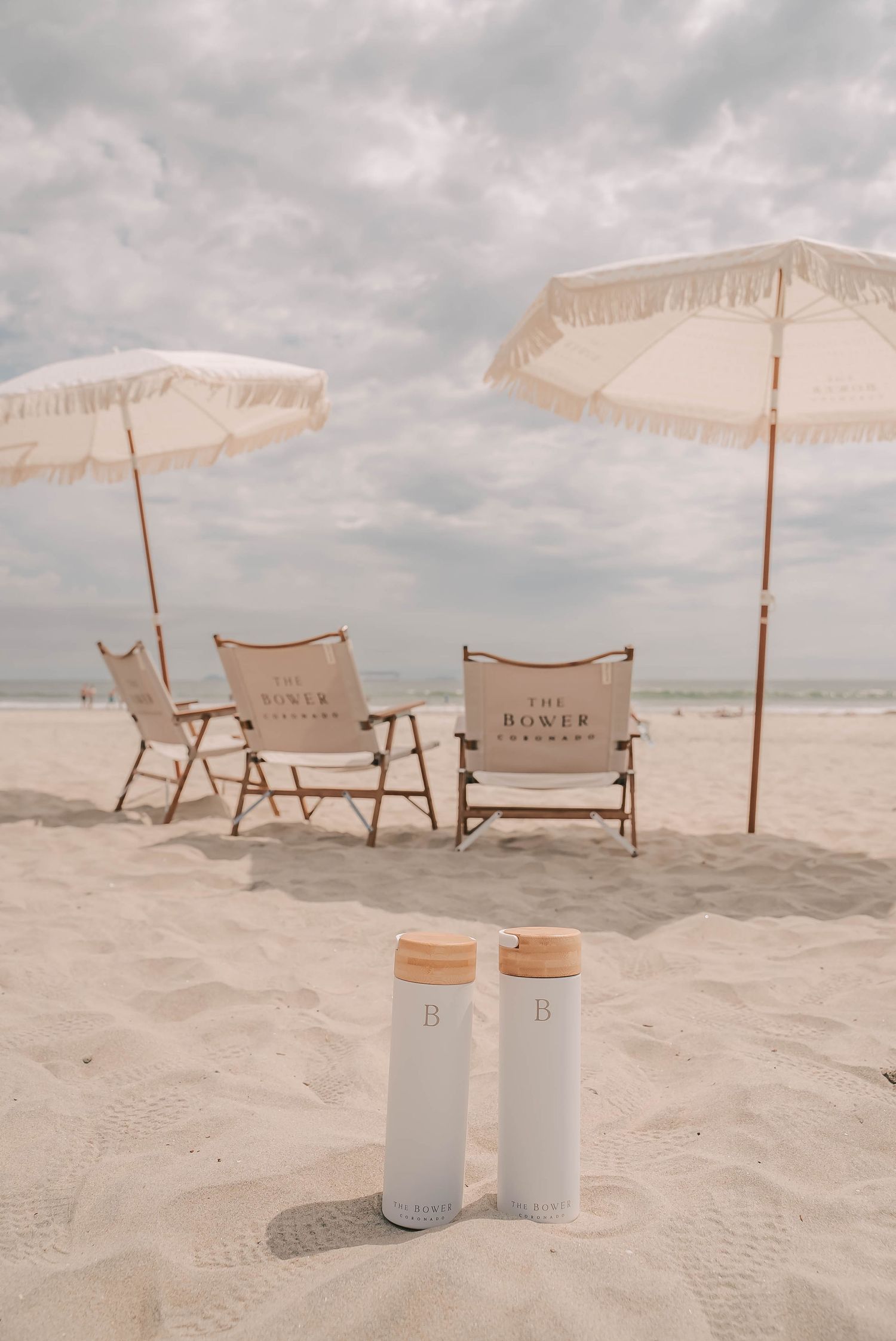 Two branded beach chairs and white fringed umbrellas on sandy shore with matching tumblers in foreground.