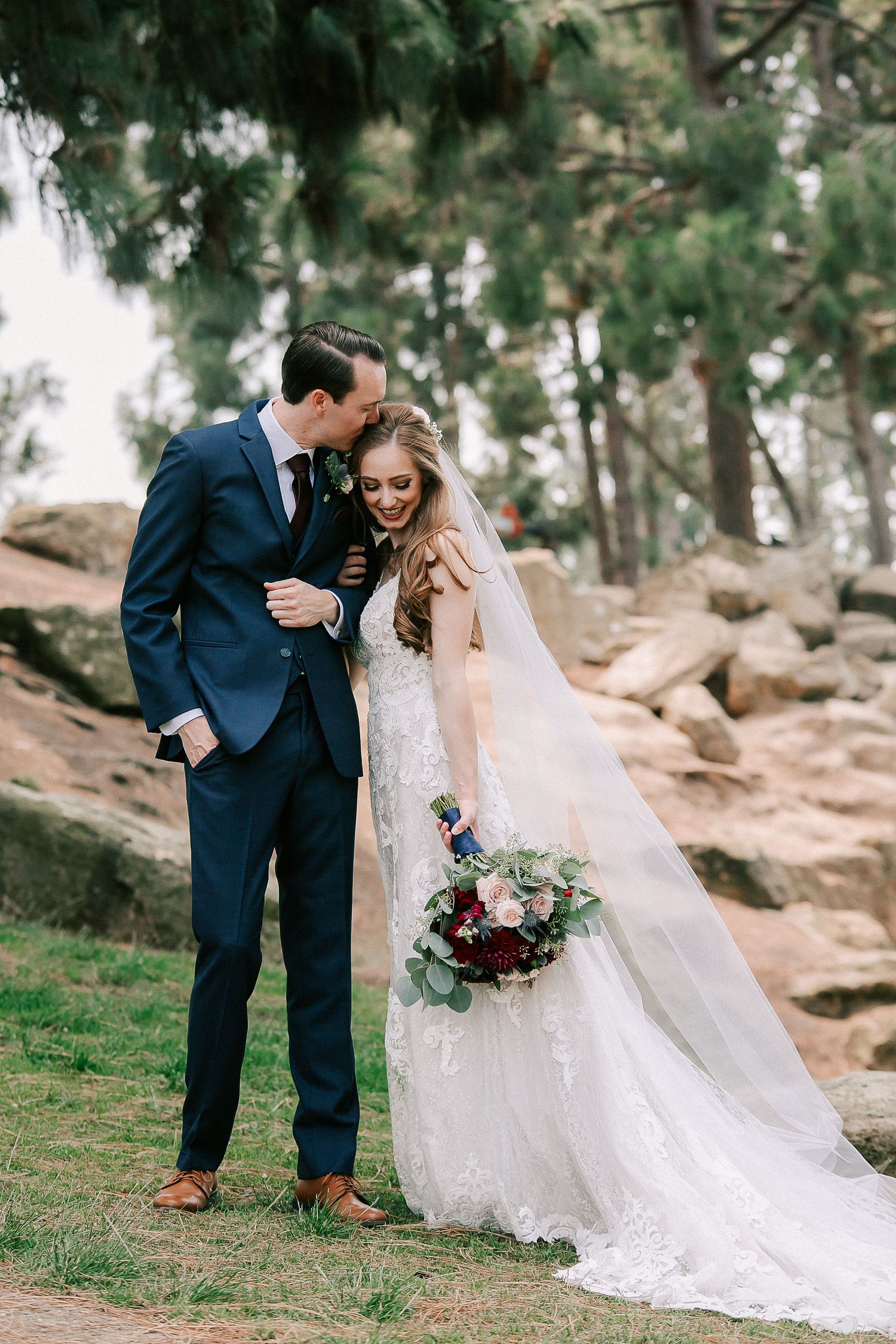 Bride and groom share a romantic moment outdoors, with lace wedding gown, veil, and bouquet of red and burgundy flowers.