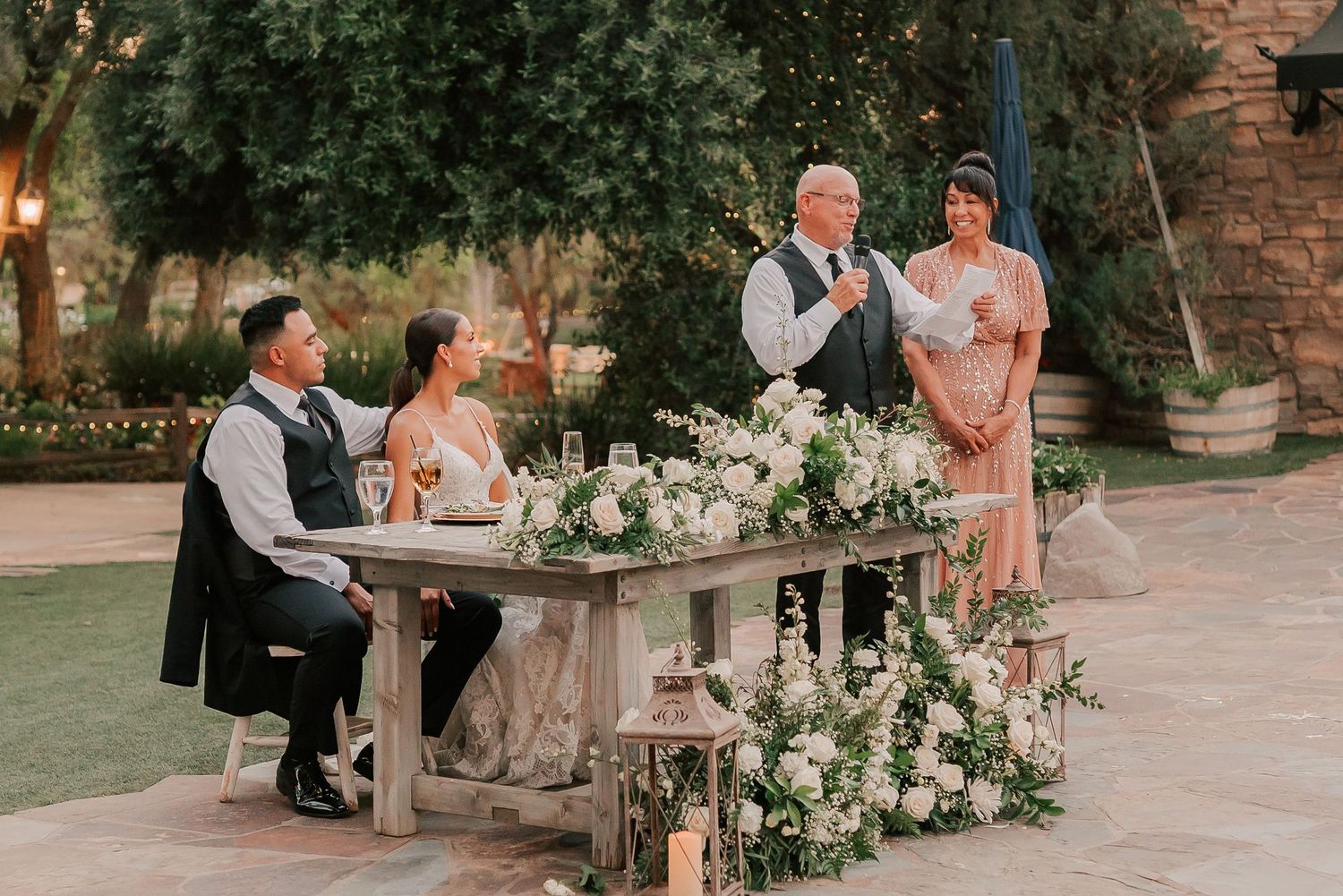 Outdoor wedding reception with couple seated at floral-decorated rustic table while officiant gives speech.