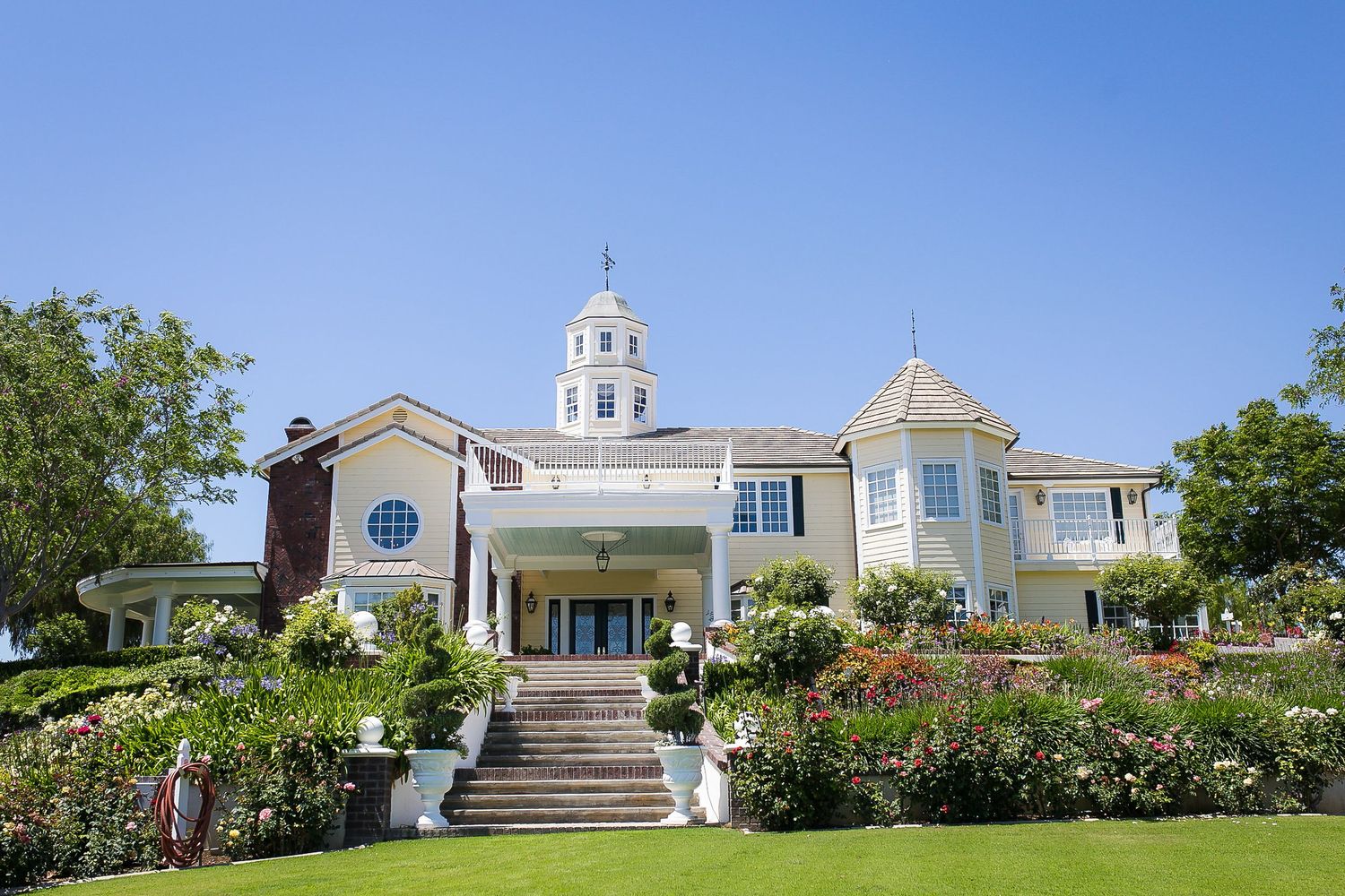 Large white colonial mansion with cupola, manicured gardens, green lawn, and colorful flowers under a clear blue sky.