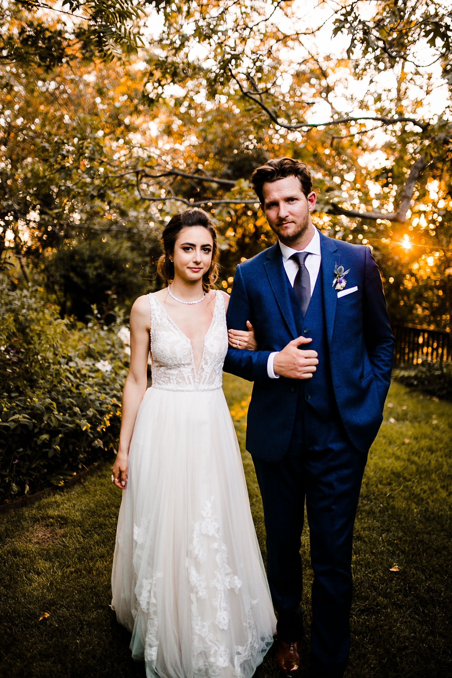 Bride in white lace gown and groom in navy suit pose together in a golden-hour garden setting.