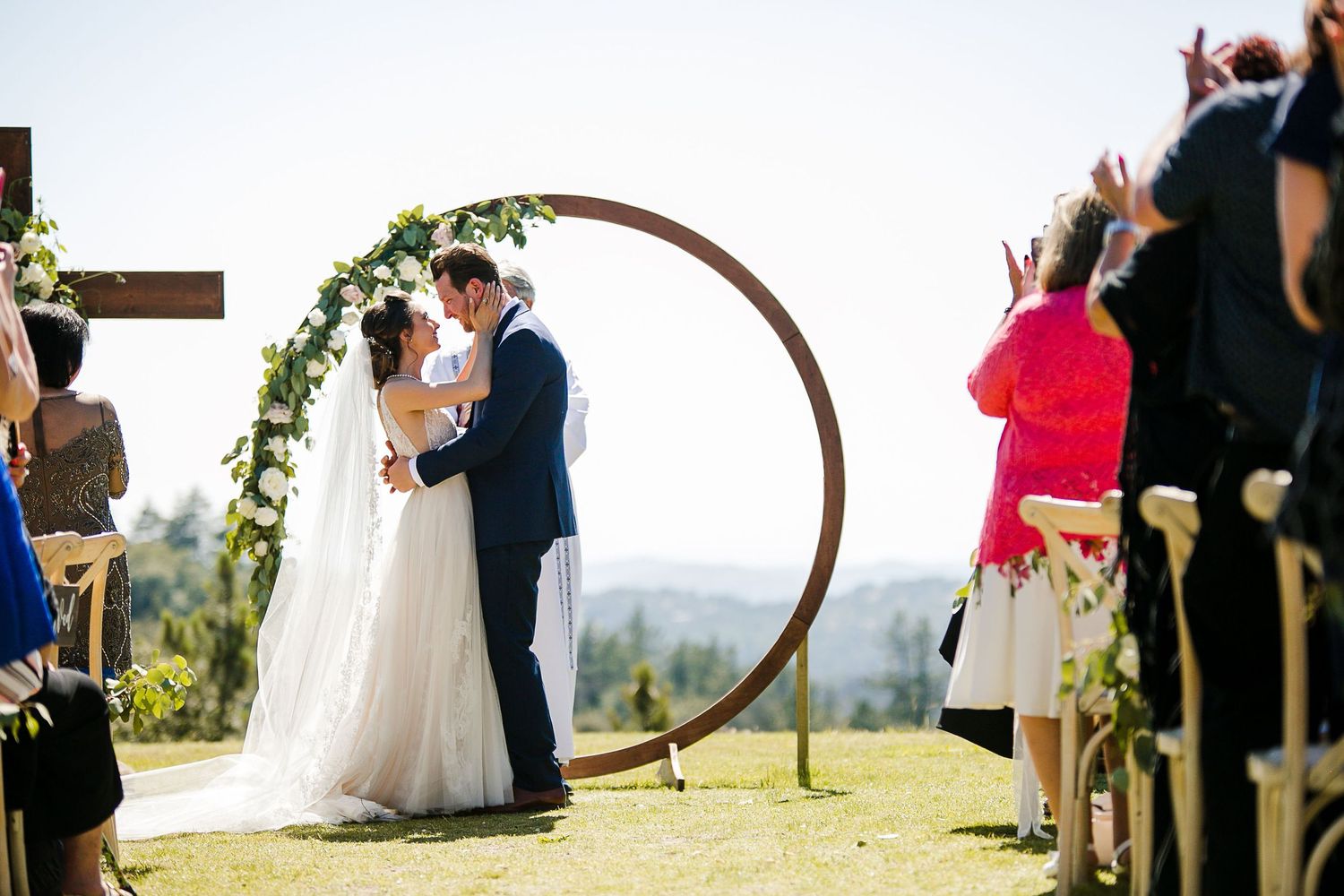 Bride and groom share first kiss under circular wooden arch adorned with greenery at outdoor wedding ceremony.