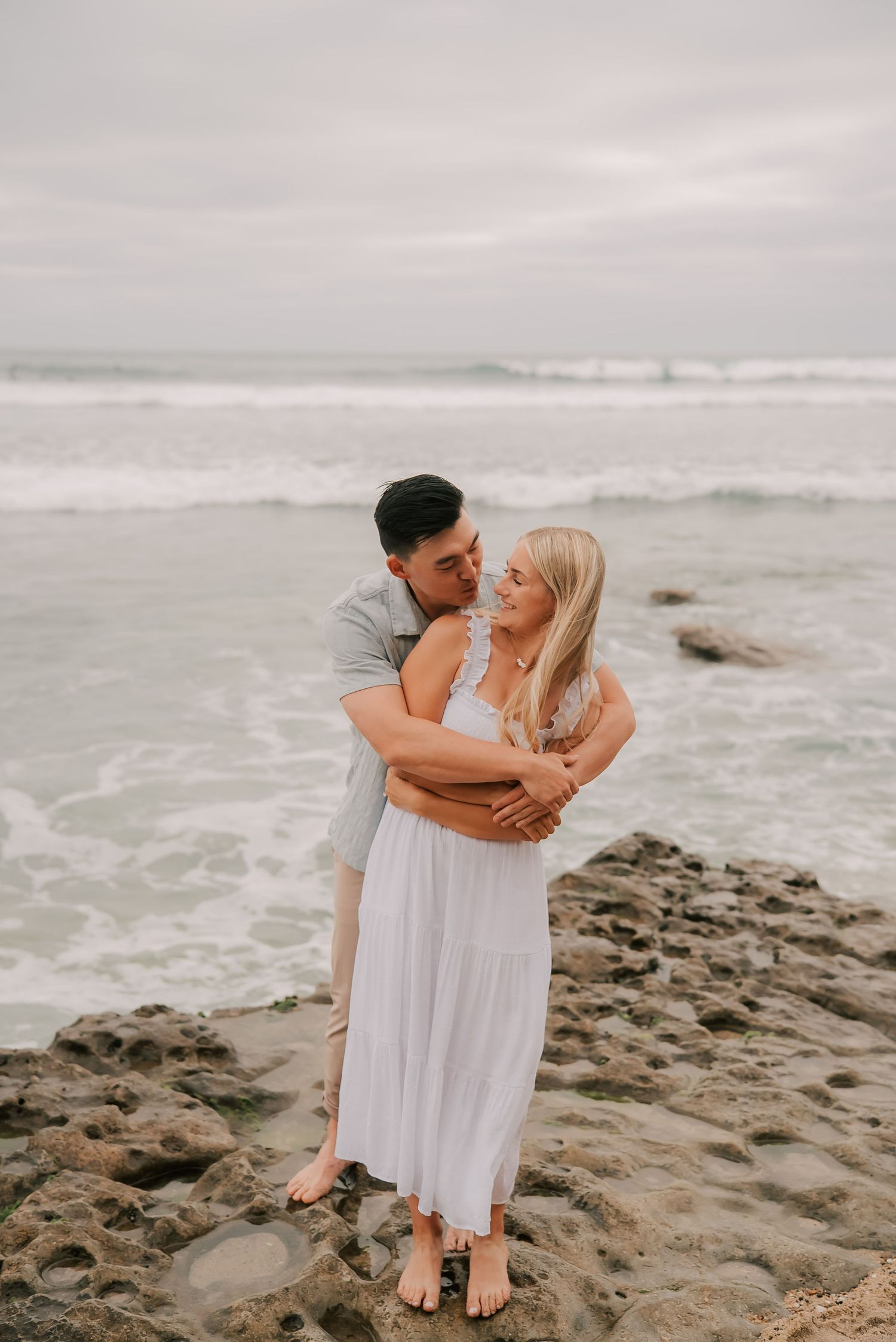 Romantic couple hugging on rocky beach, woman in white maxi dress, with ocean waves behind them.