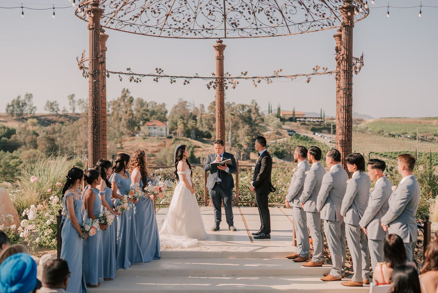 Outdoor wedding ceremony under ornate pergola with bridesmaids in blue dresses and groomsmen in gray suits, scenic vineyard backdrop.