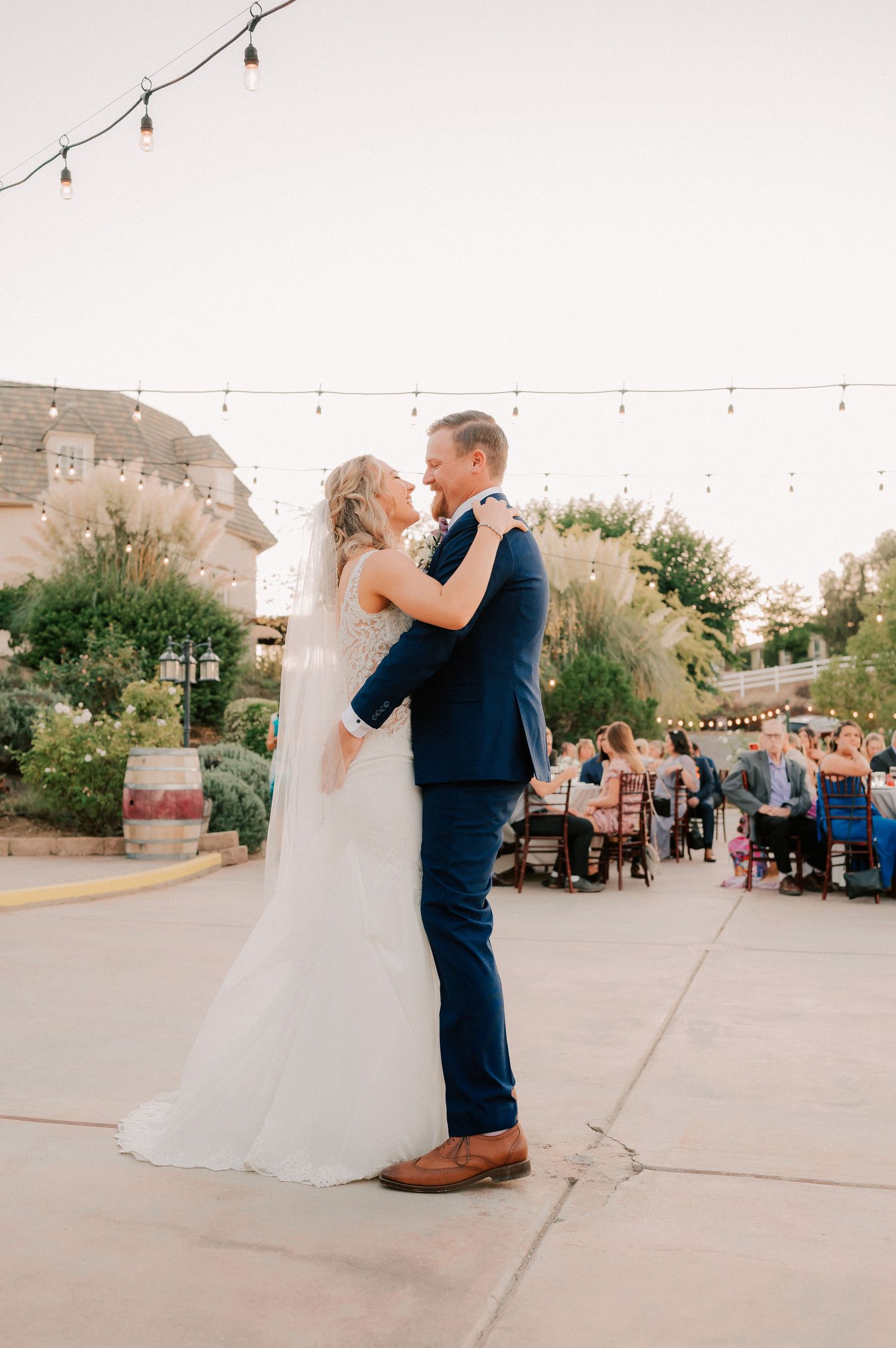 Bride and groom share first dance outdoors at wedding reception with string lights and guests seated in background.