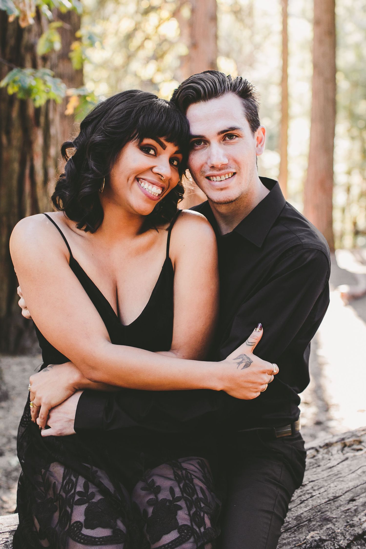 A smiling couple in black outfits embrace warmly while sitting outdoors among trees in a forest setting.