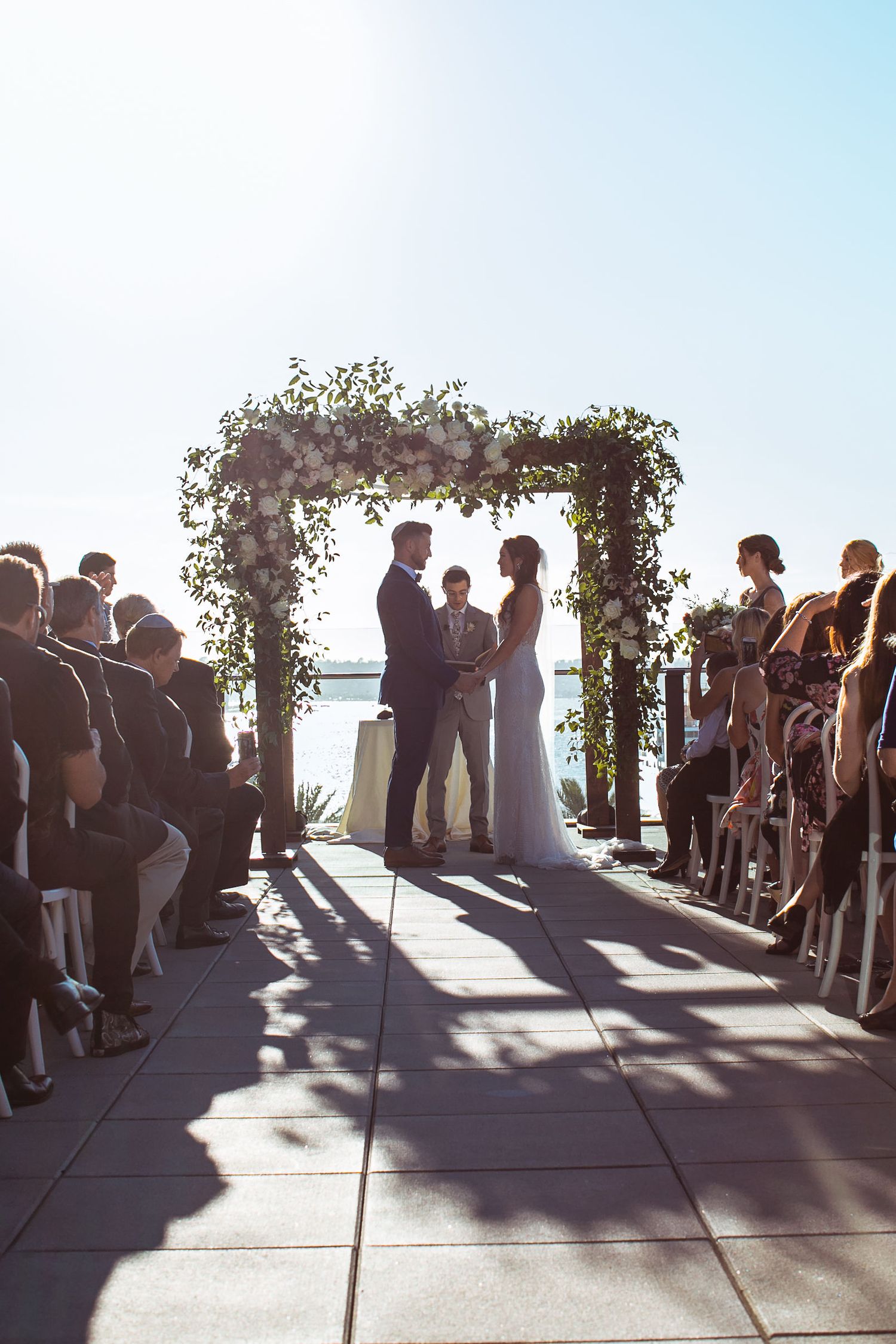 Couple exchanging vows under floral arch at outdoor waterfront wedding ceremony with seated guests lining the aisle.