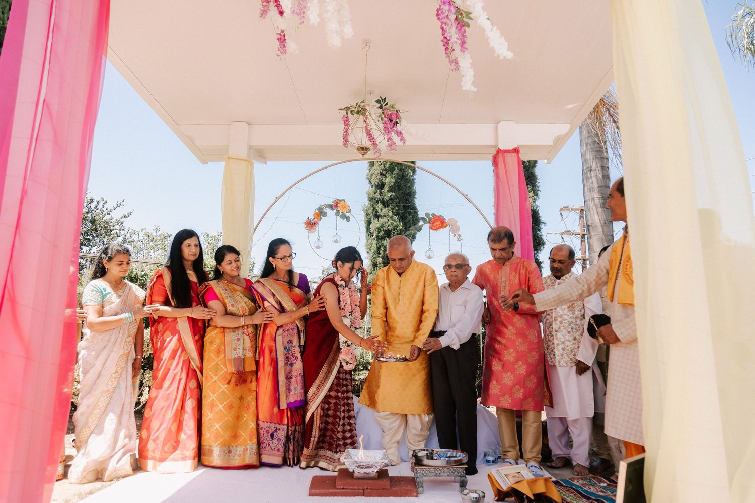Group of guests in colorful traditional Indian attire gathered under a decorated mandap during a wedding ceremony.