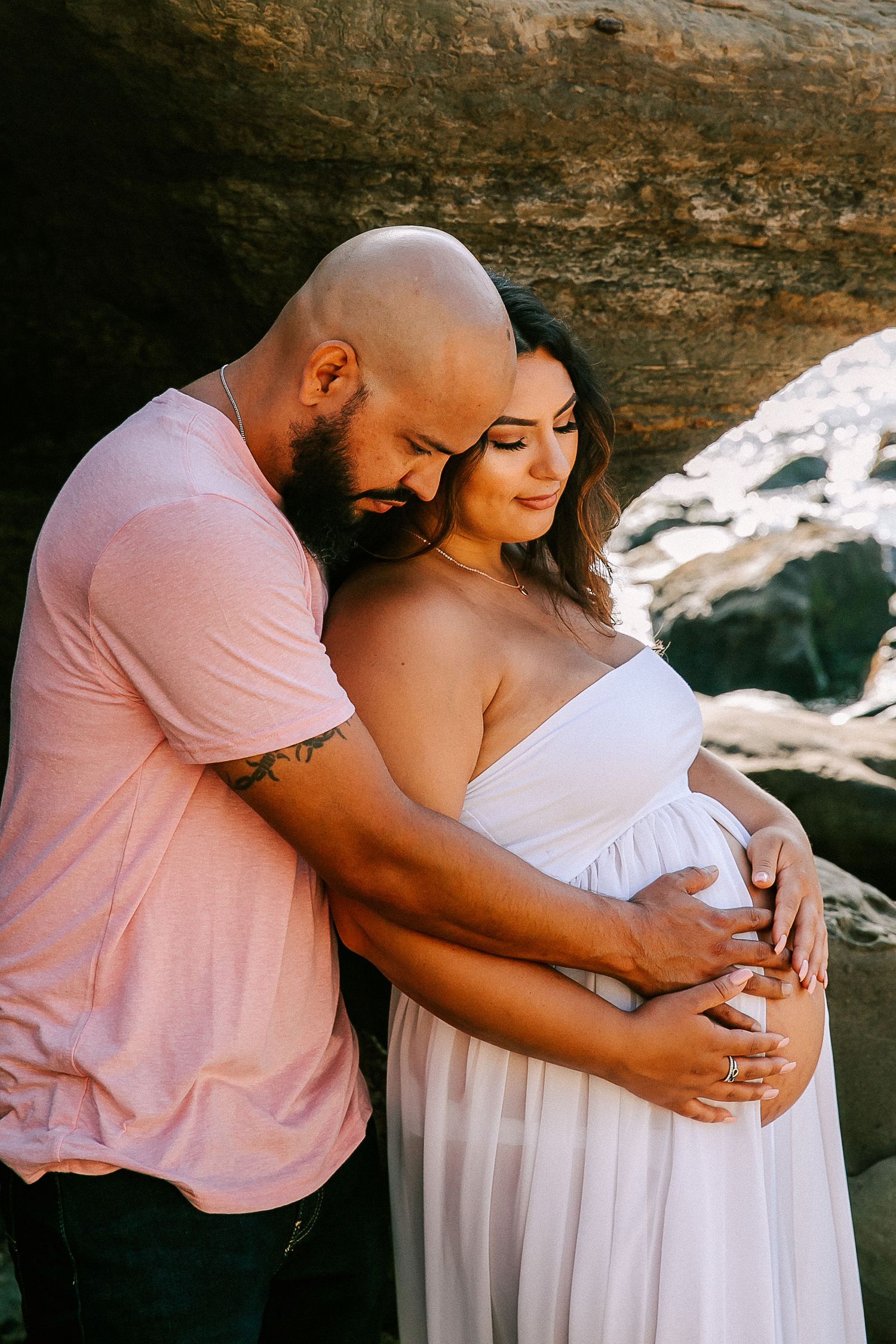 Couple embracing under rock arch, man holding pregnant woman in white gown from behind tenderly.