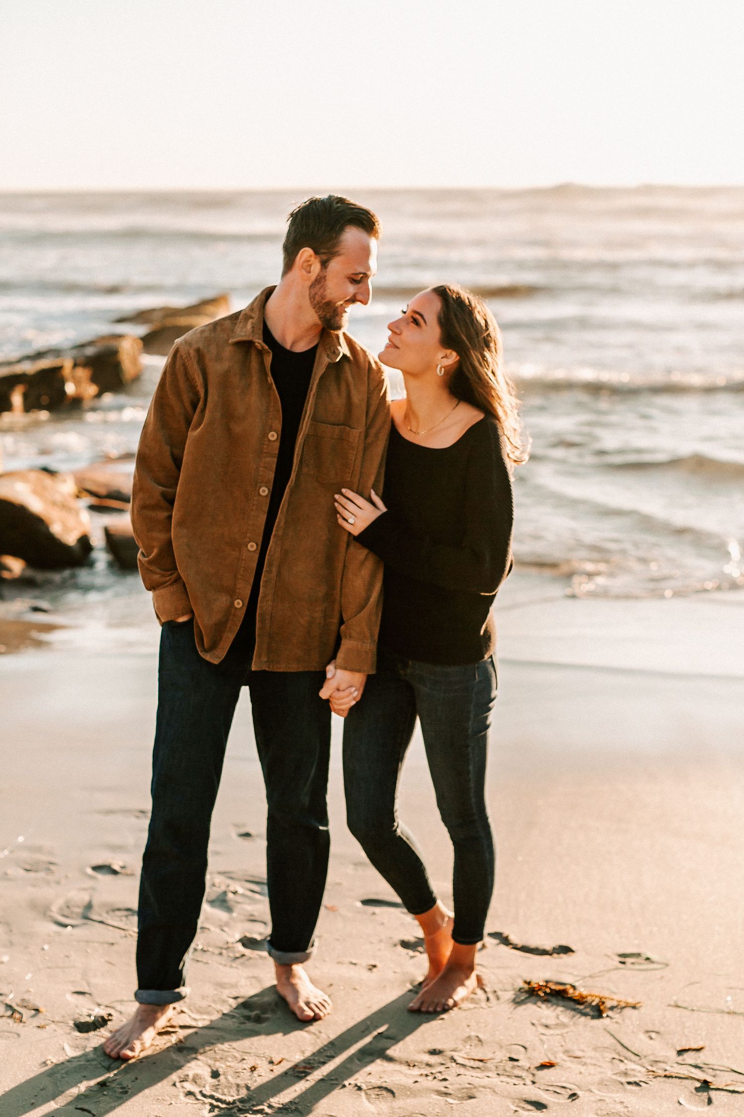 Couple walking hand in hand on a sunny beach, gazing at each other with the ocean behind them.